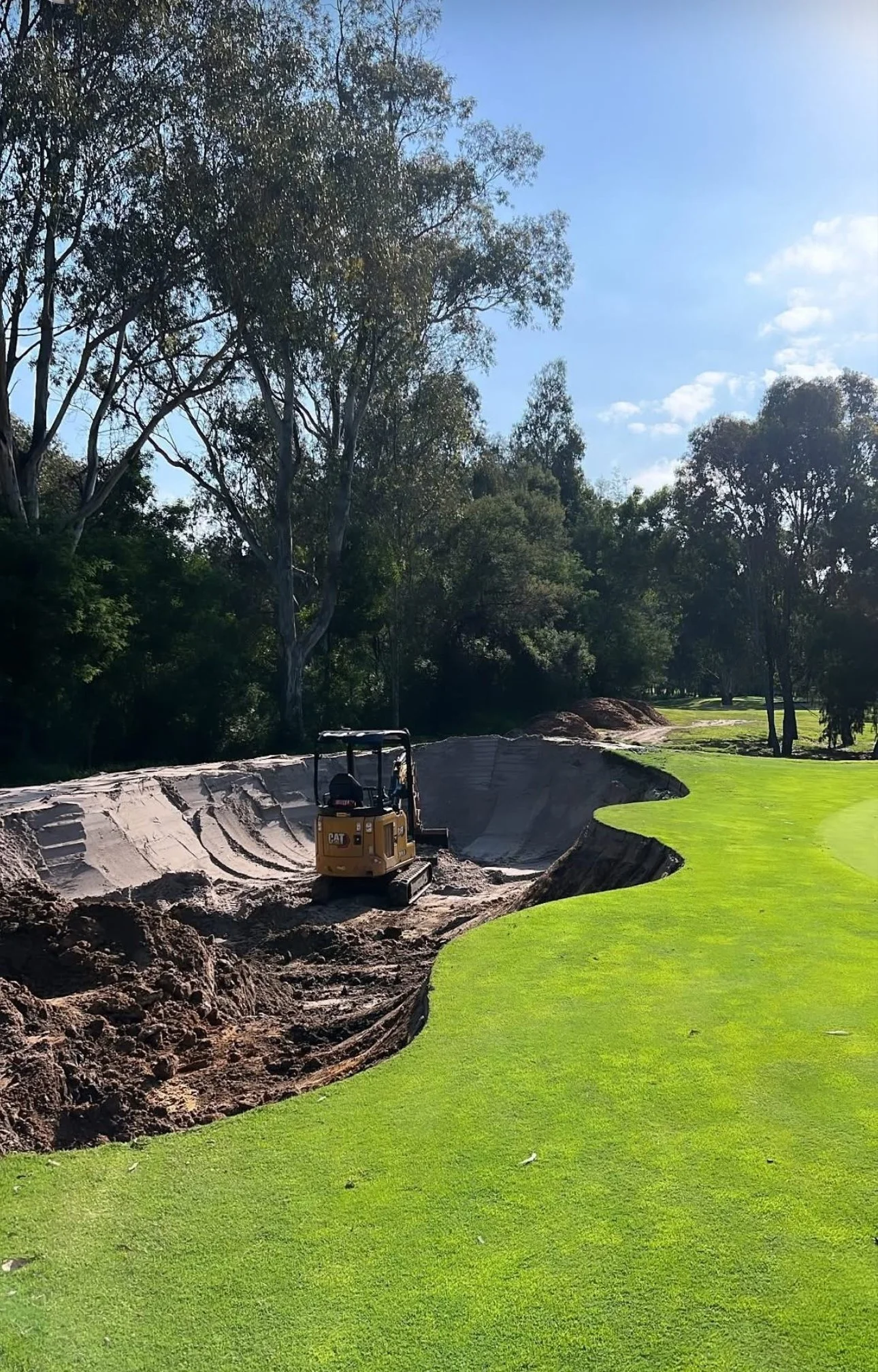 A small yellow Caterpillar excavator working on a golf course, with a dirt bunker and green grass, surrounded by trees under a partly cloudy sky.