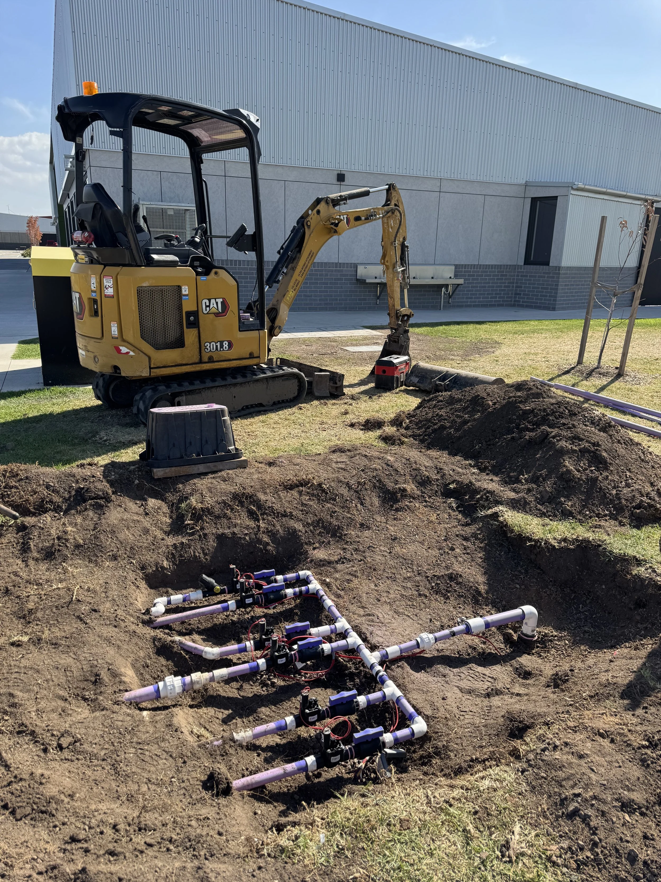Construction site with a small yellow CAT excavator working on a plumbing installation, with pipes and valves in the dug-up ground outside a building.