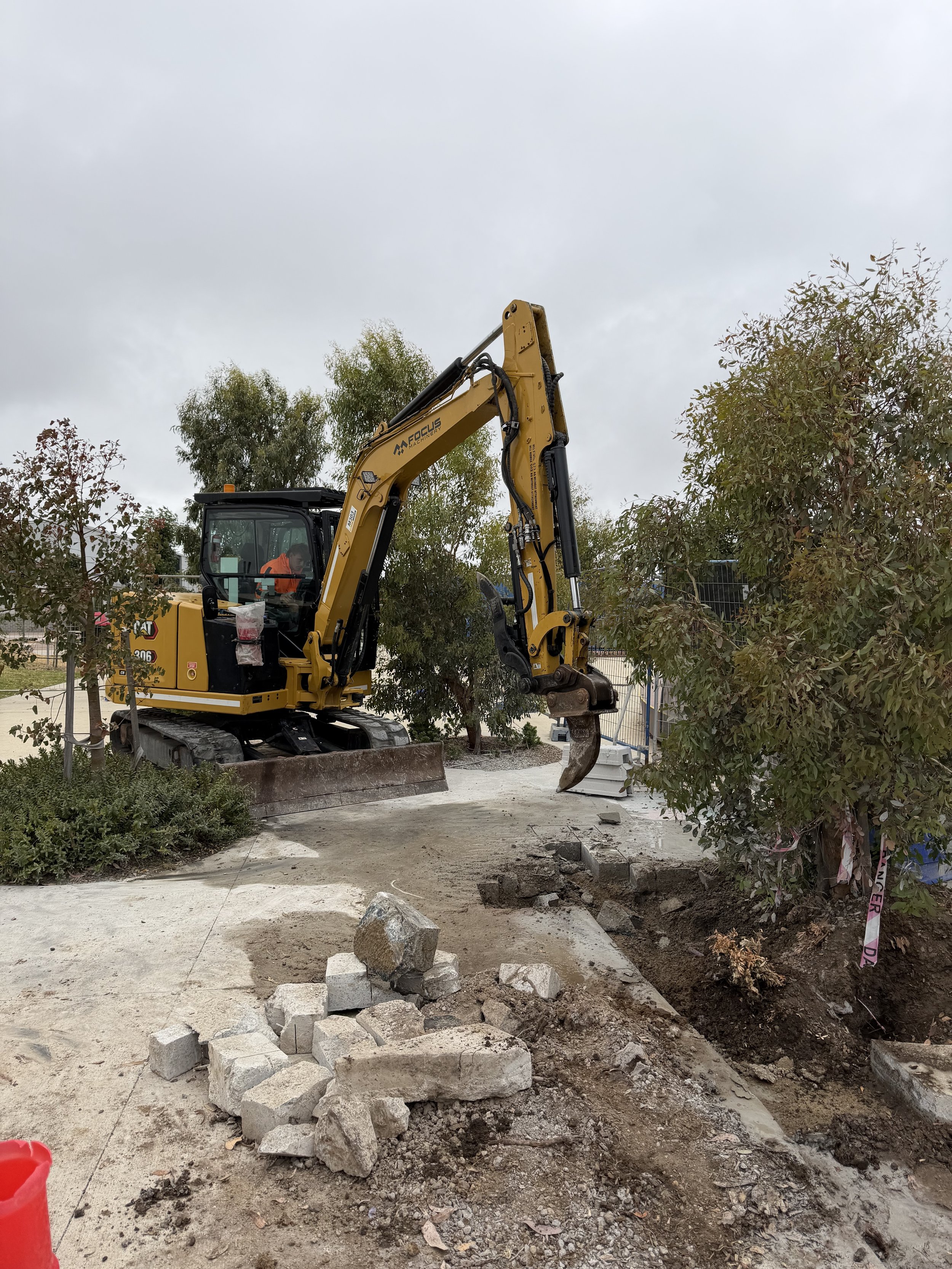 A construction excavator is digging near trees and a pathway with scattered rocks and dirt, under a cloudy sky.