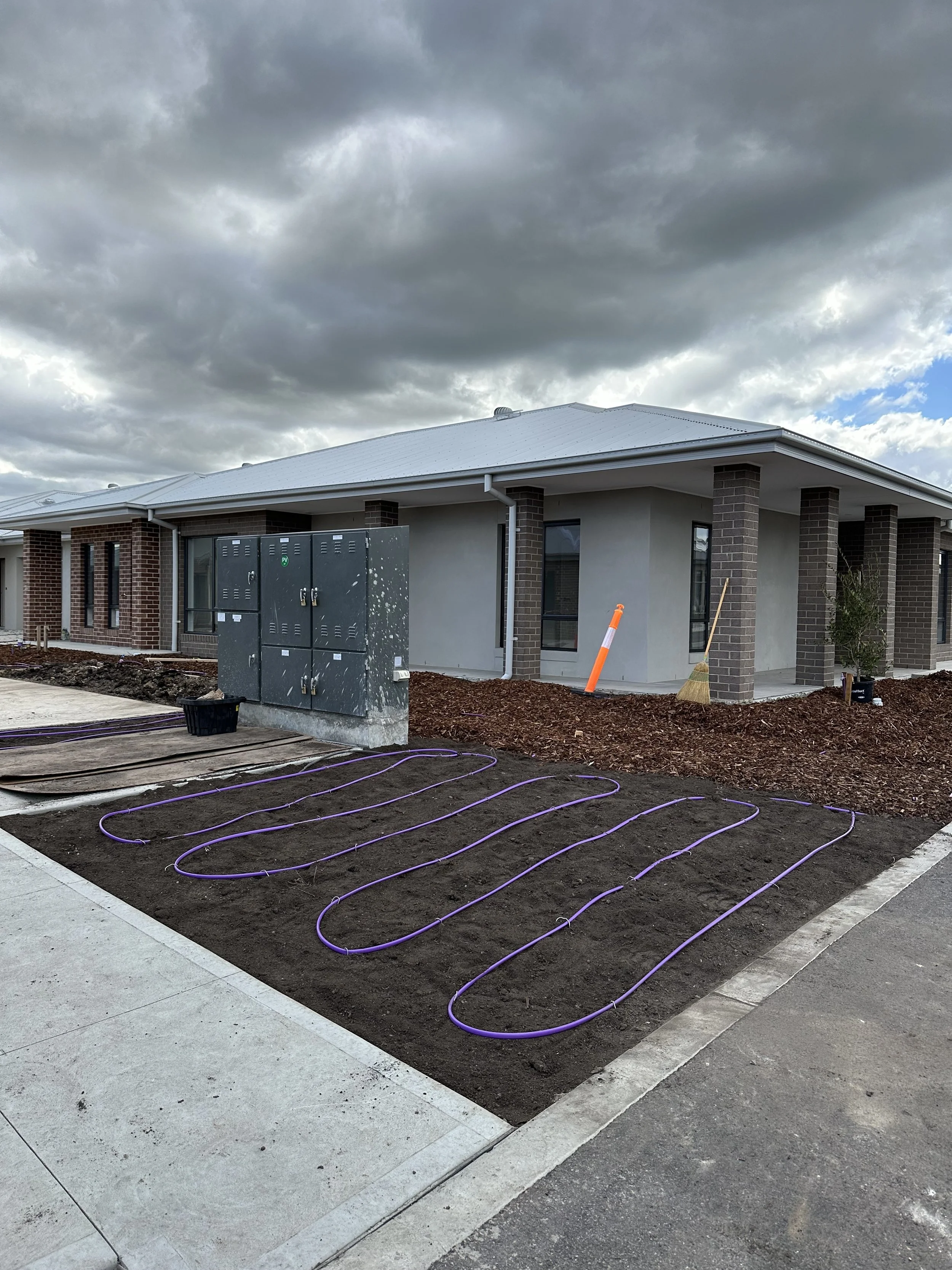 Construction site with an unfinished house, under cloudy skies, with purple tubing in the ground for landscaping or irrigation installation.