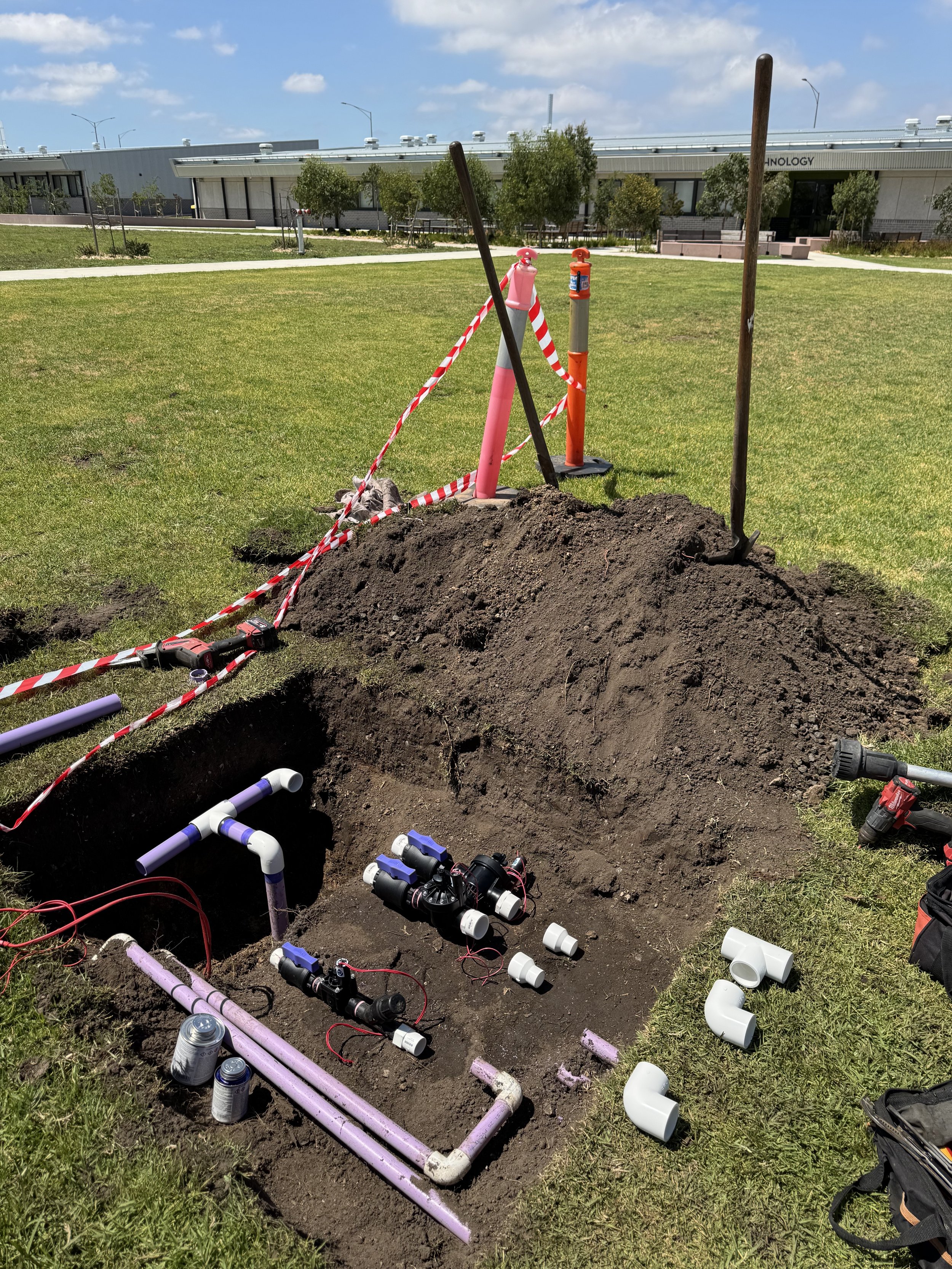 Installation of underground piping with multiple PVC pipes, valves, and connections in a grass field, sectioned off with safety barriers.