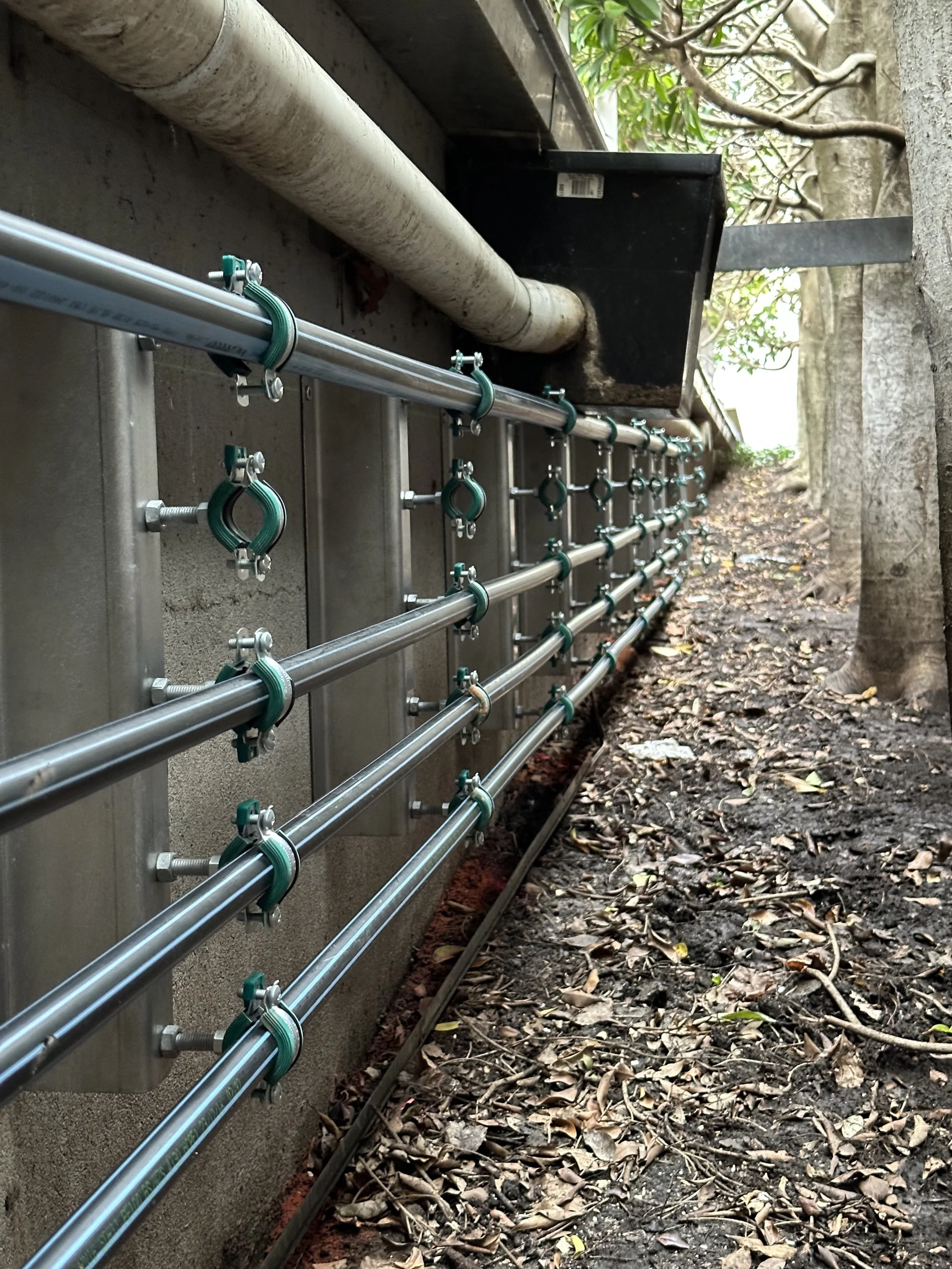 Close-up view of metal pipes and green fittings attached to a wall outside. The pipes are secured with green clamps, and there's a white pipe along the top. The setting appears to be a garden or outdoor area with trees and dirt ground.