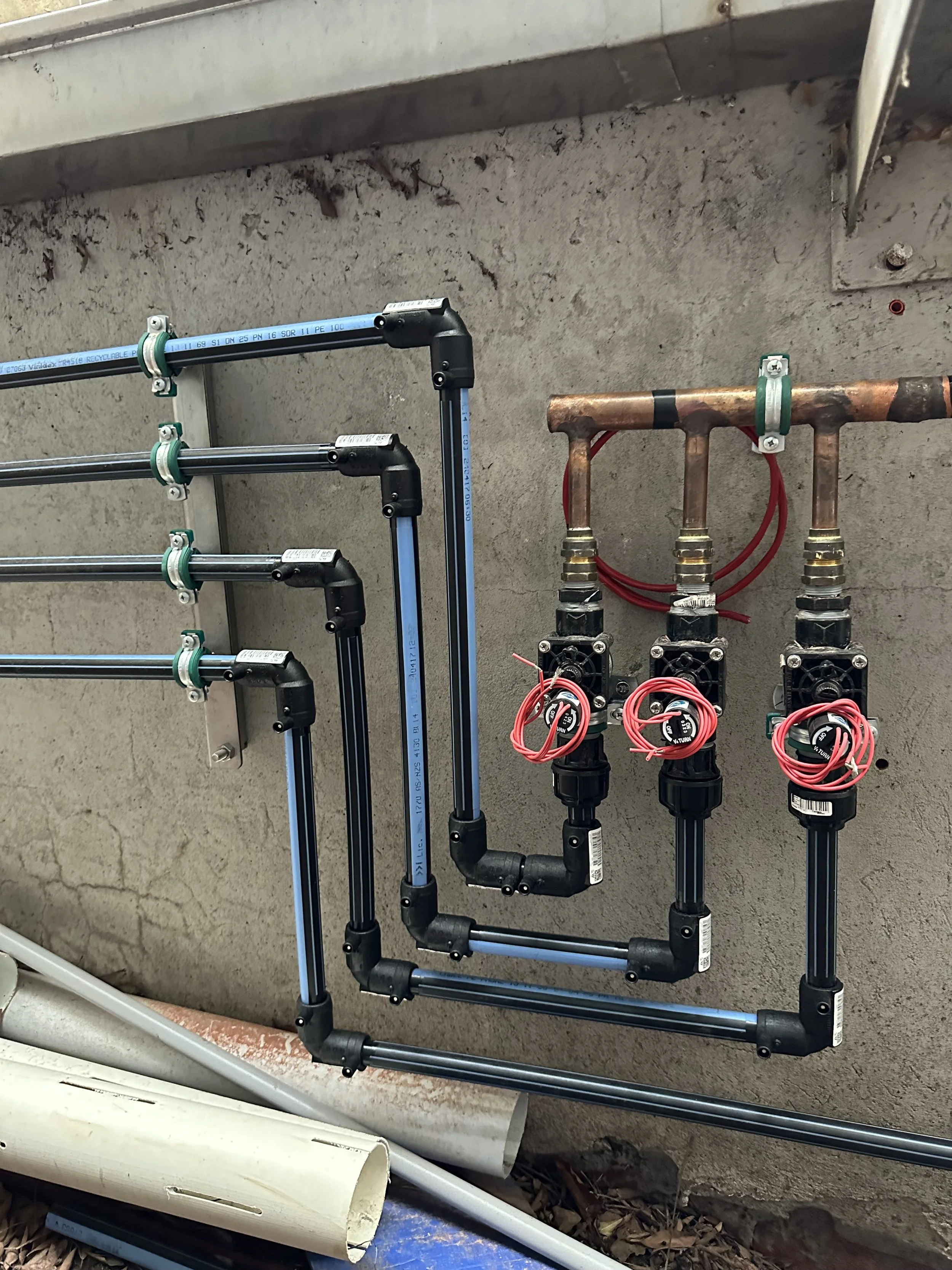Plumbing system with copper pipes, black valves, and red tubing connected on a concrete wall, with blue PEX pipes and fittings, and some rolled white material on the ground nearby.