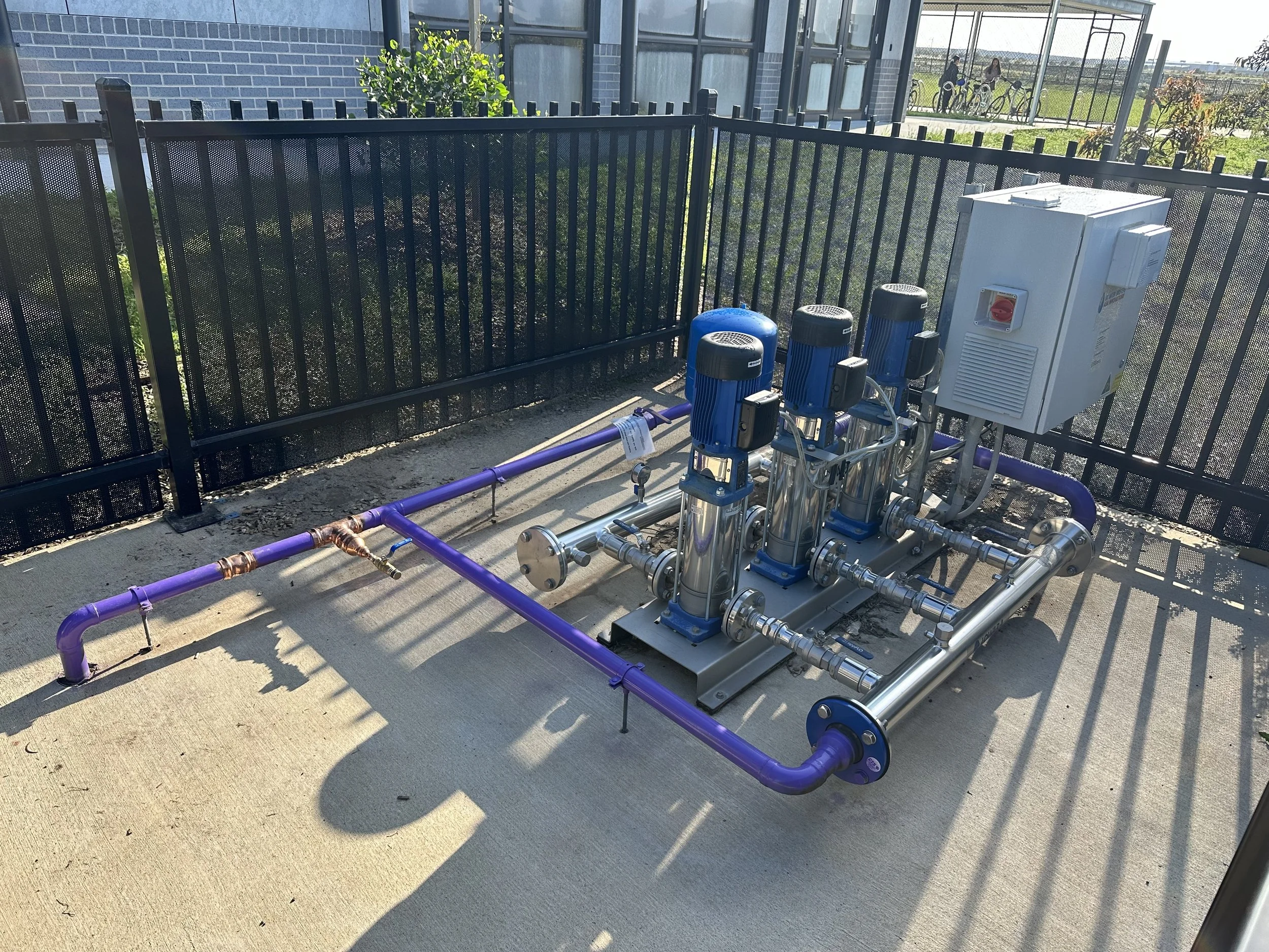 Industrial water pump system with three vertical blue motorized pumps, purple pipes, and a white control box installed outdoors on a concrete pad, fenced with black metal fencing, with a greenhouse and bicycles in the background.