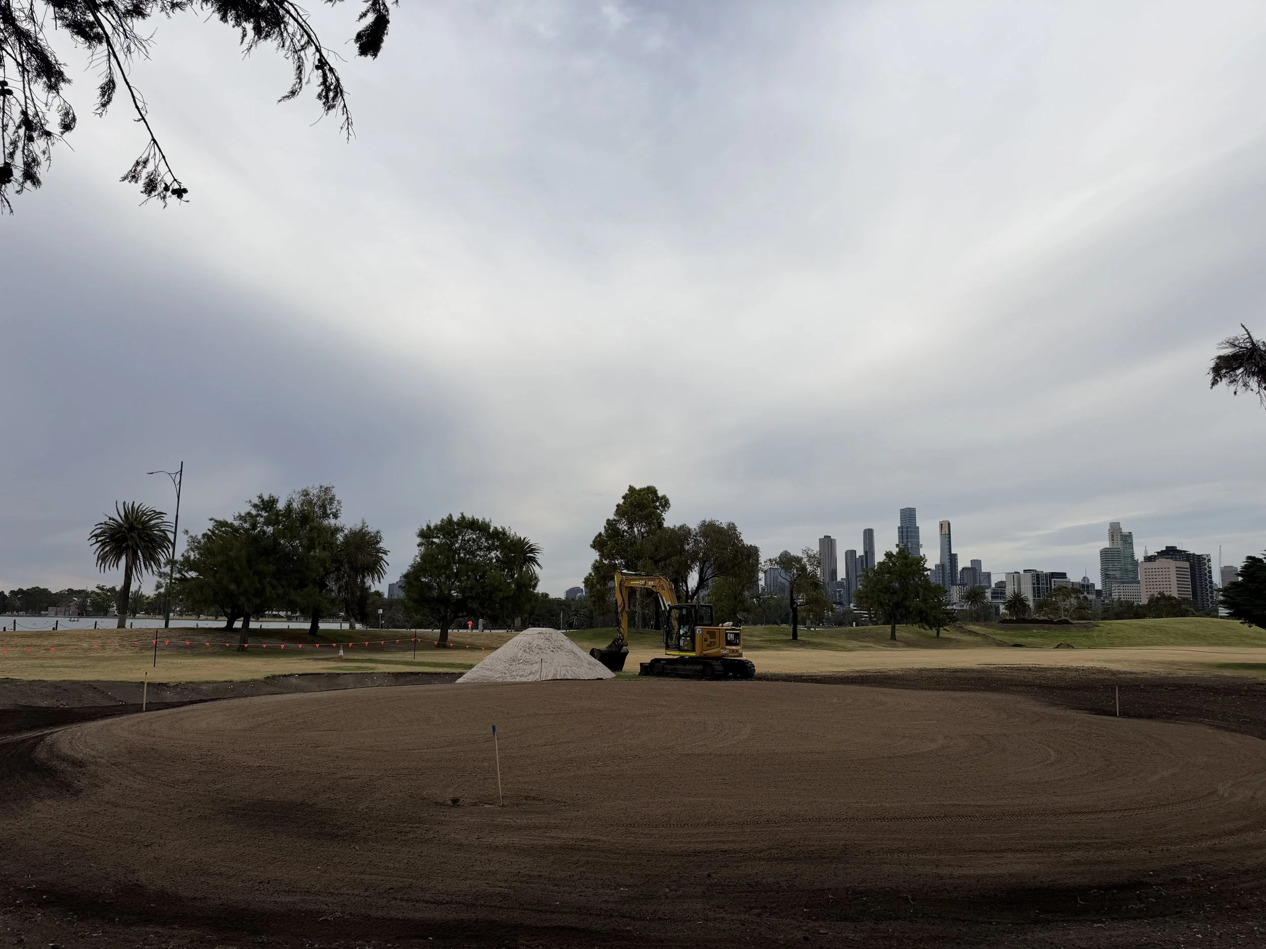 Construction site with a small yellow excavator and piles of gravel in a park, with city skyline in the background and overcast sky.