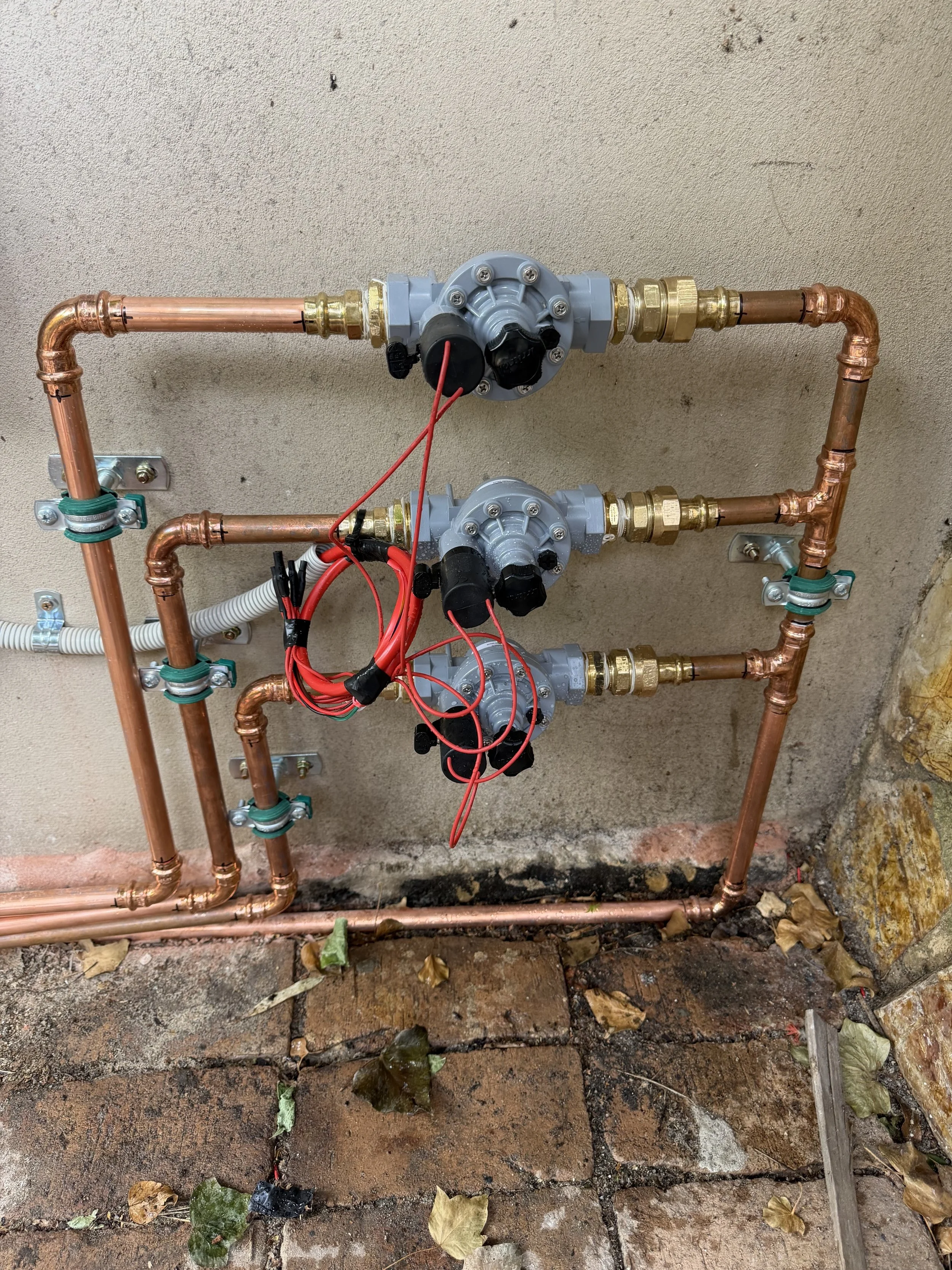 A plumbing system with three copper water meters and valves, with red wires connected to the meters, mounted on a concrete wall outside on a brick surface, surrounded by fallen leaves.