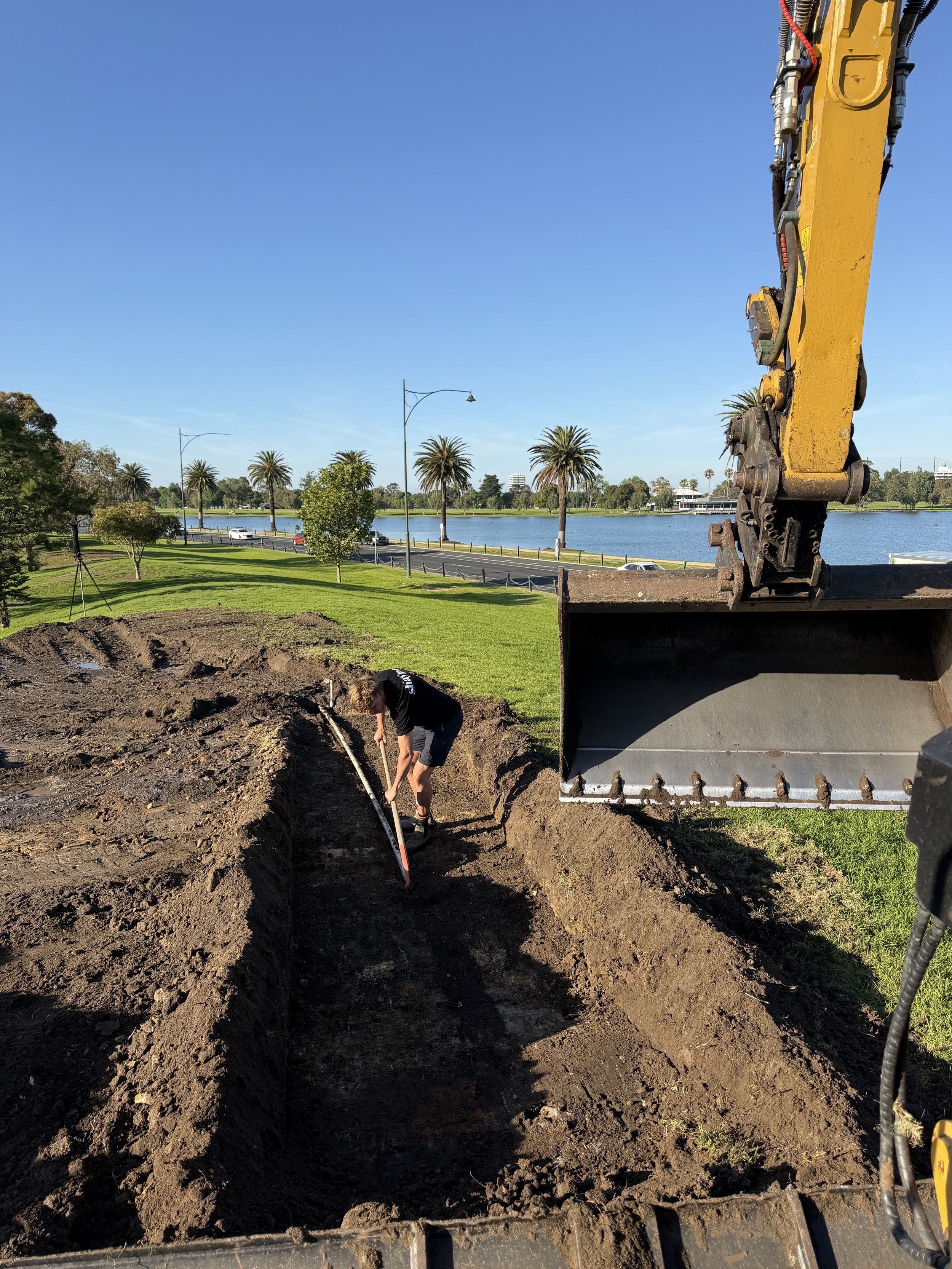 Person digging a trench with a measuring tape near construction equipment by a lake with palm trees and clear blue sky.