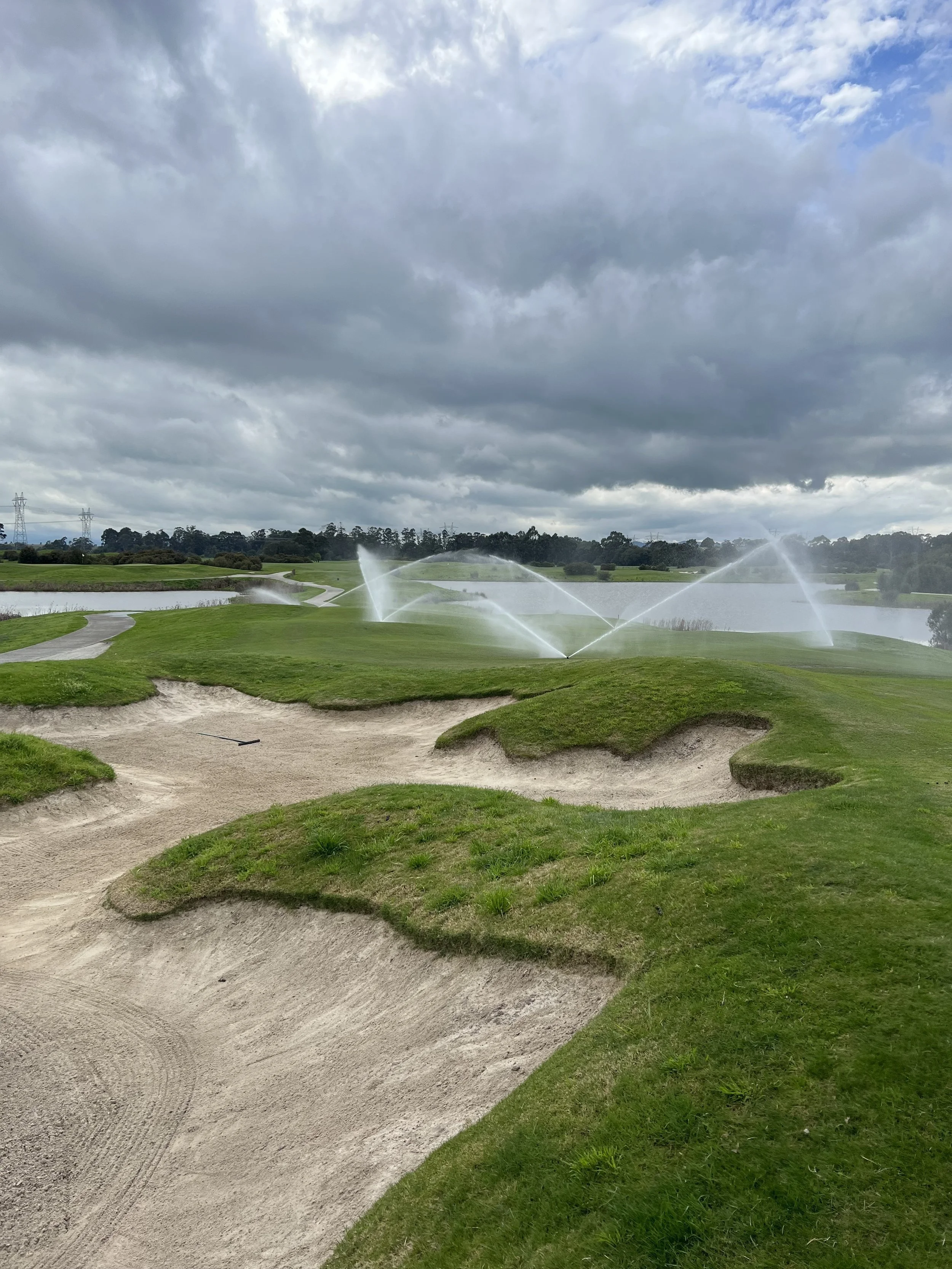 A golf course with sand bunkers, green grass, and irrigation sprinklers watering the course under a cloudy sky.