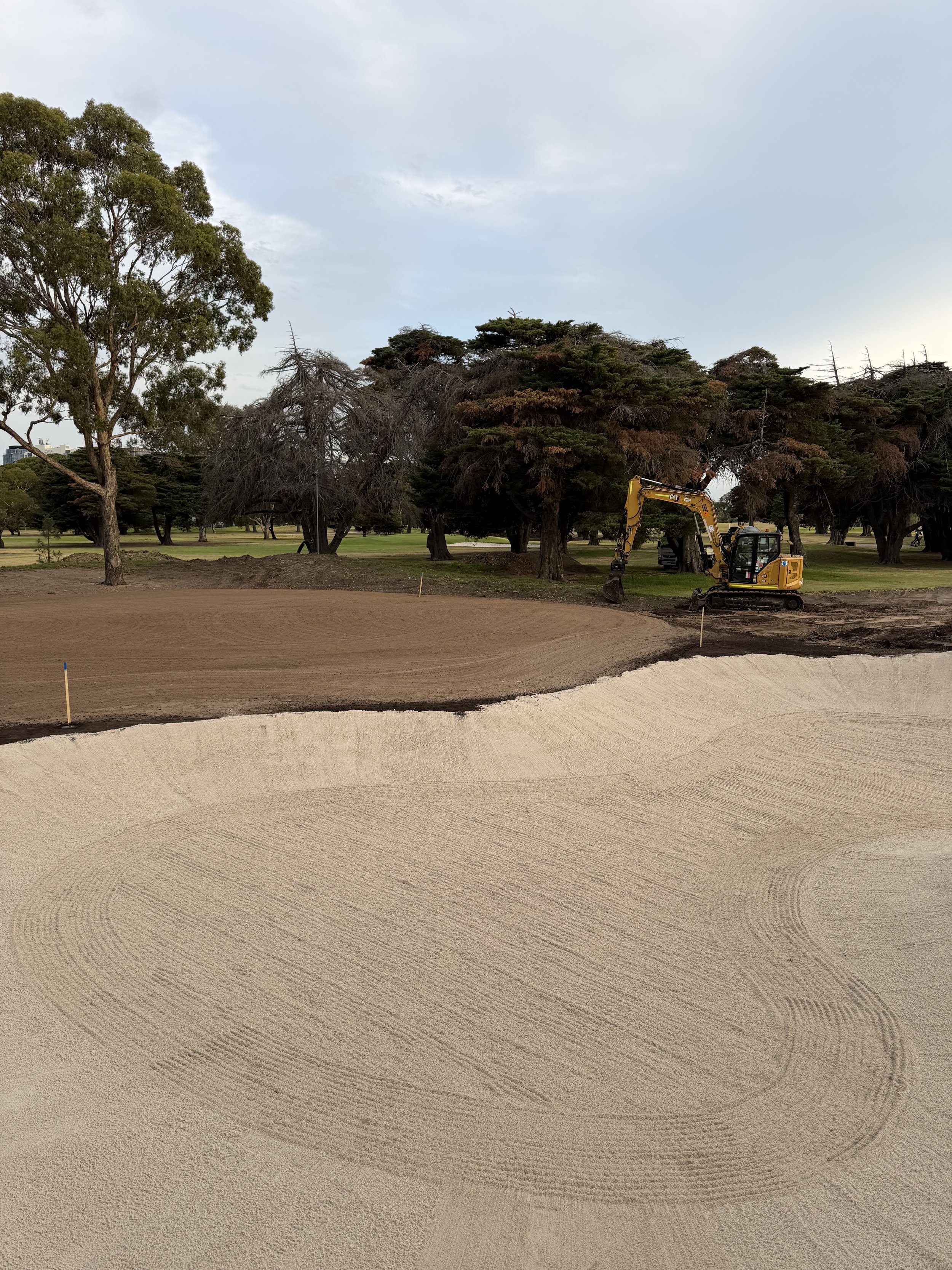A construction site on a golf course where a small yellow excavator is working near large trees, with a sand bunker in the foreground and a cloudy sky overhead.