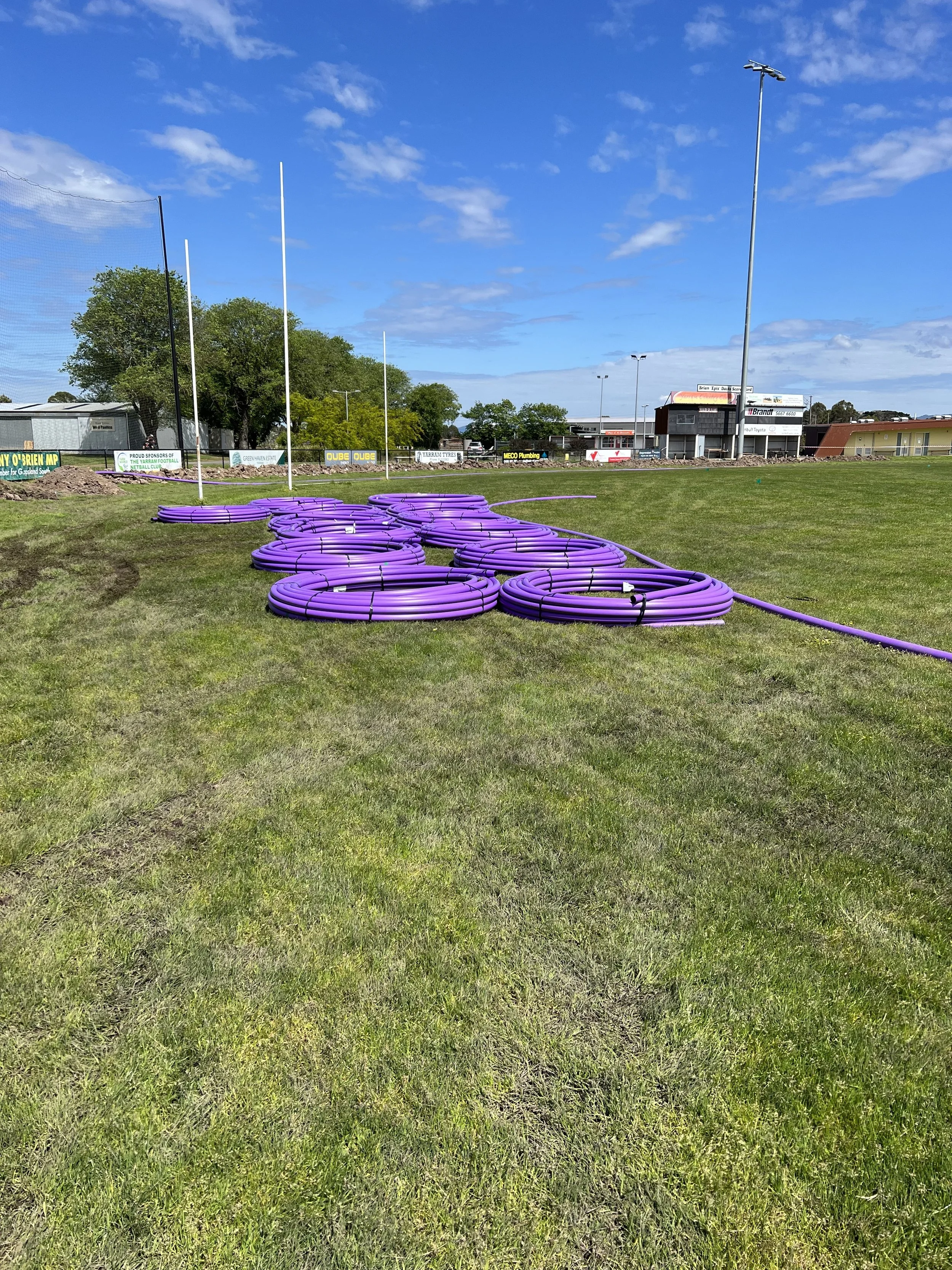Purple hoses coiled on grass in a sports field with goal posts, trees, light poles, and buildings in the background under a partly cloudy sky.