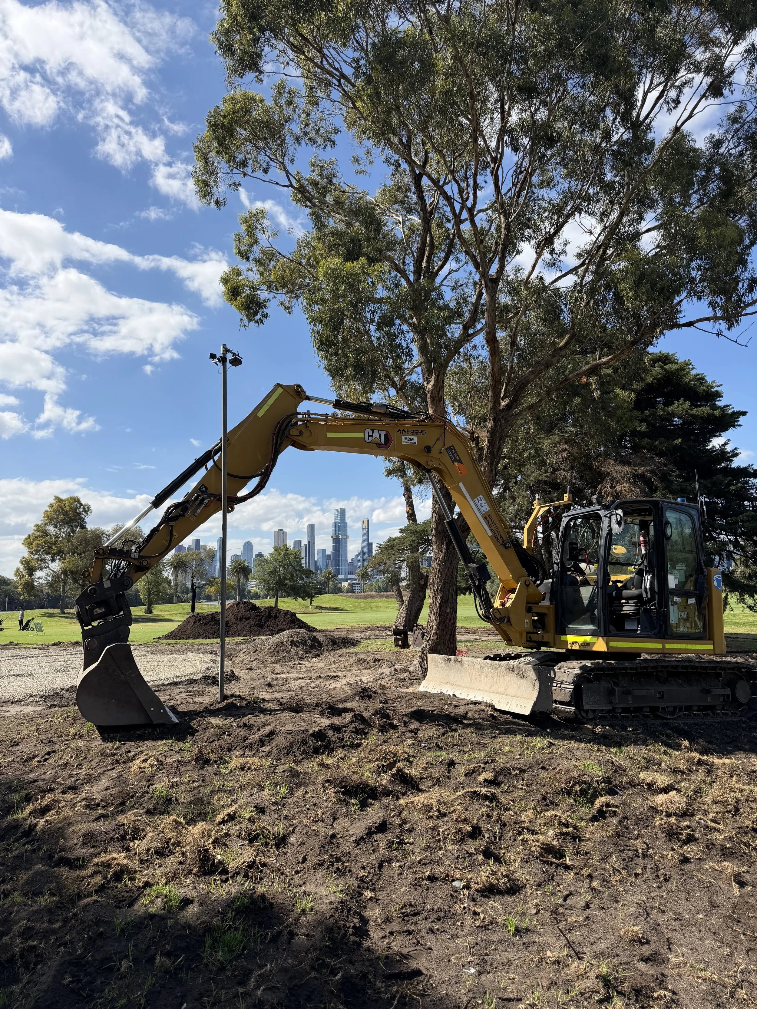 A yellow excavator digging into the ground in a park with trees, a city skyline, and a blue sky with clouds in the background.
