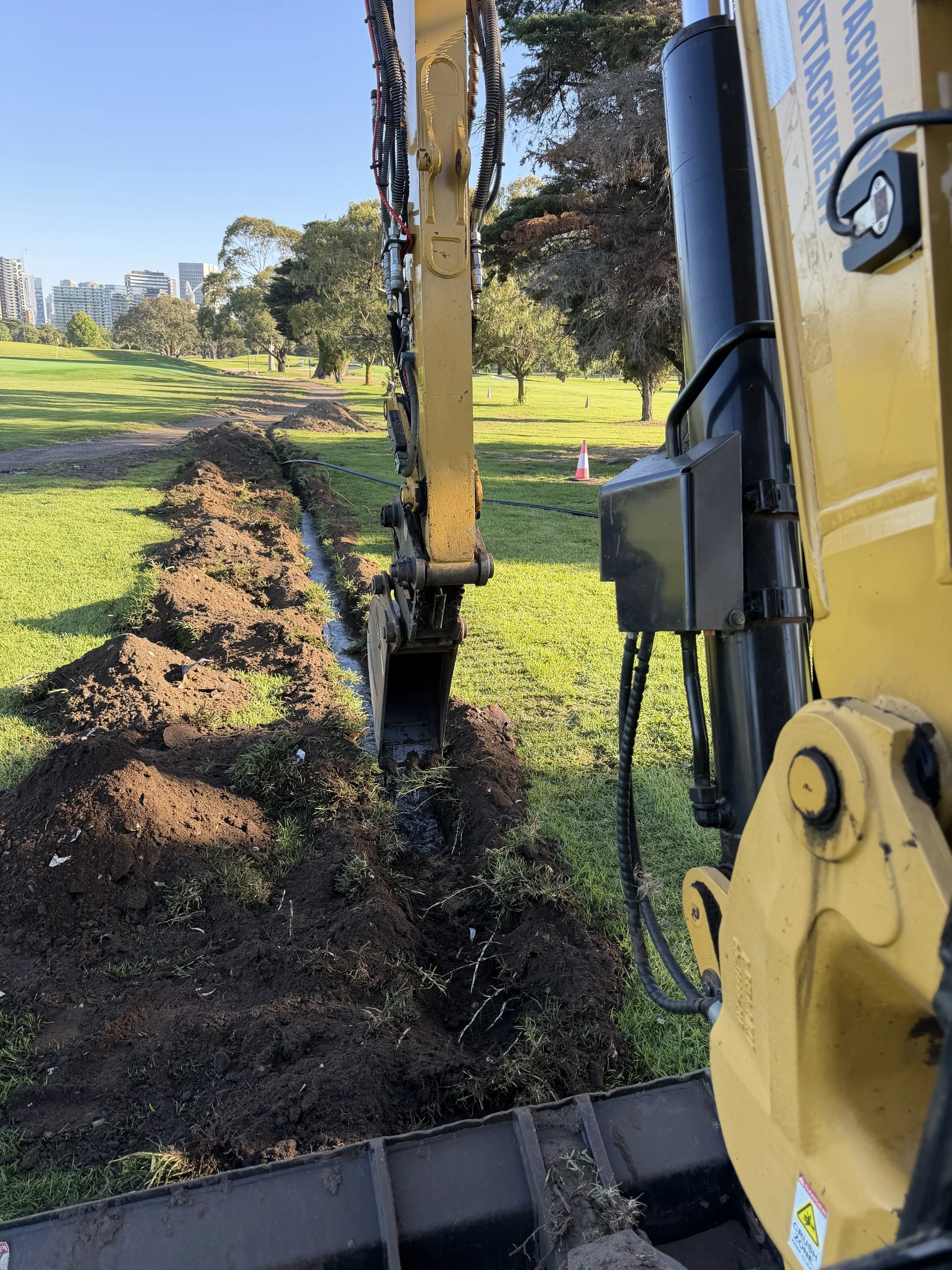 A yellow excavator digging a trench in a grassy park with trees and city buildings in the background.