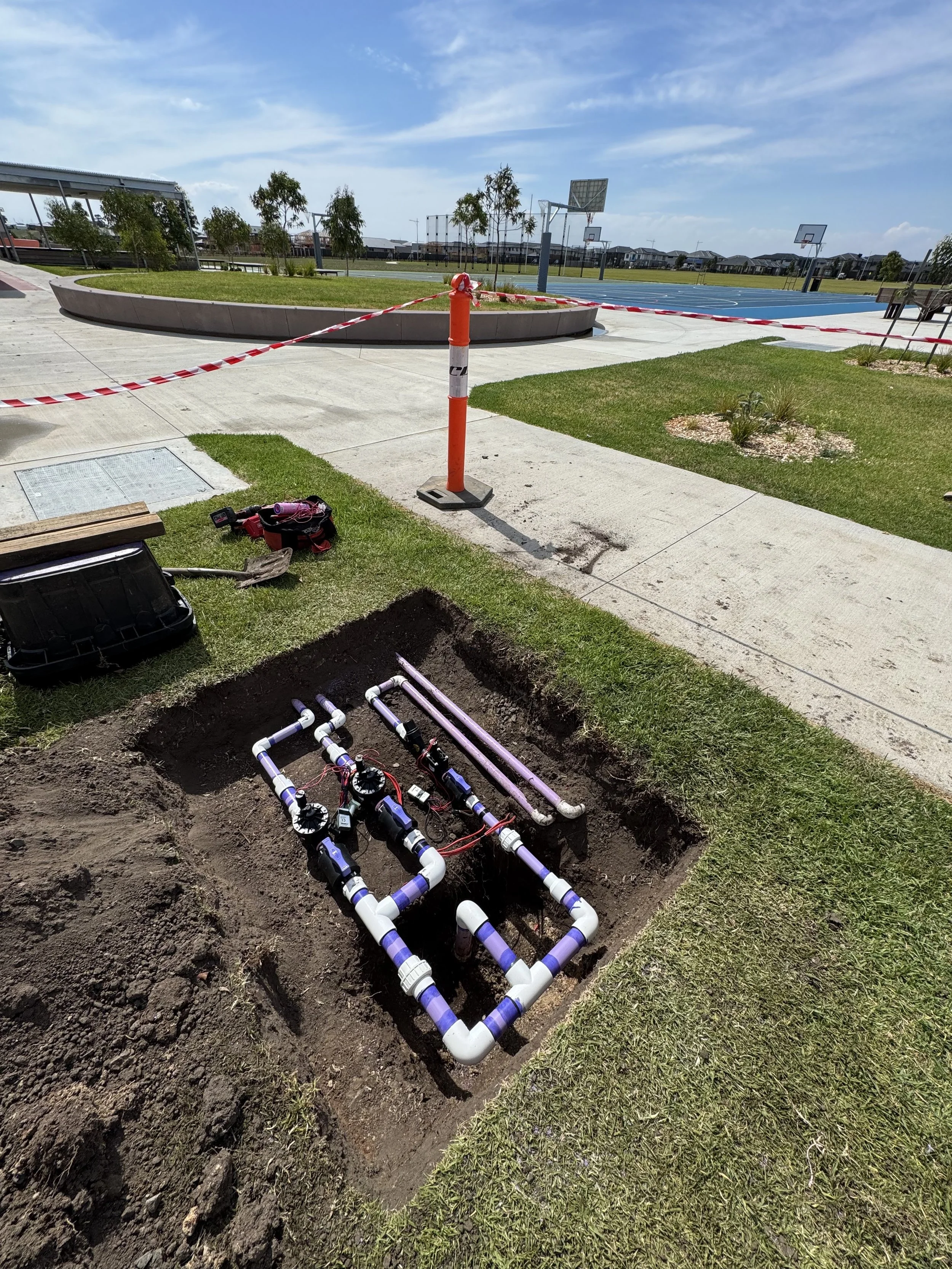 Underground plumbing system installation with white and purple pipes, valves, and fittings in a dug trench on a grassy area near a sidewalk, with a construction barrier and sports courts in the background.
