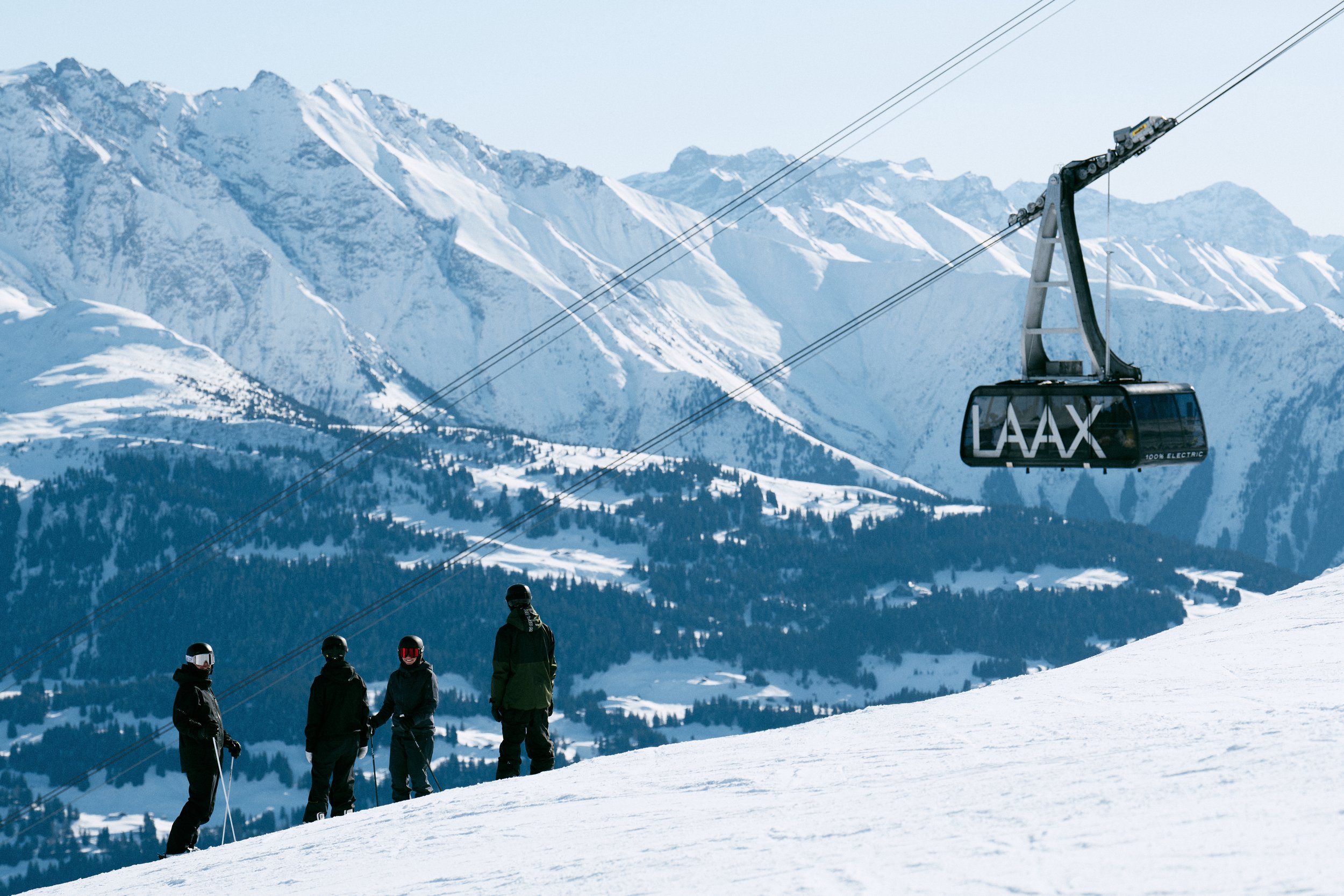 Four people wearing ski gear standing on a snowy mountain slope, with a ski lift and snowy mountains in the background.