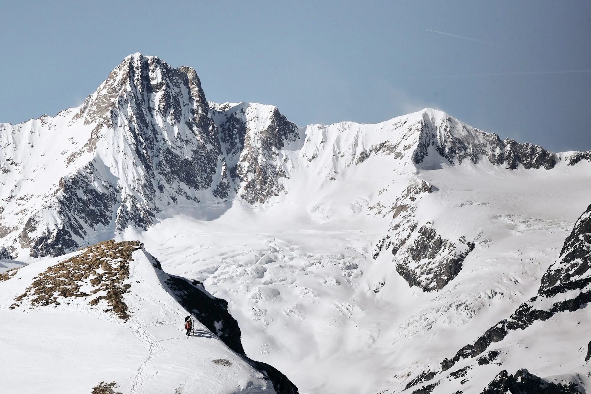 Snow-covered mountain peaks with some rocky patches, a glacier, and two climbers with backpacks and trekking poles ascending a snow slope in the foreground.