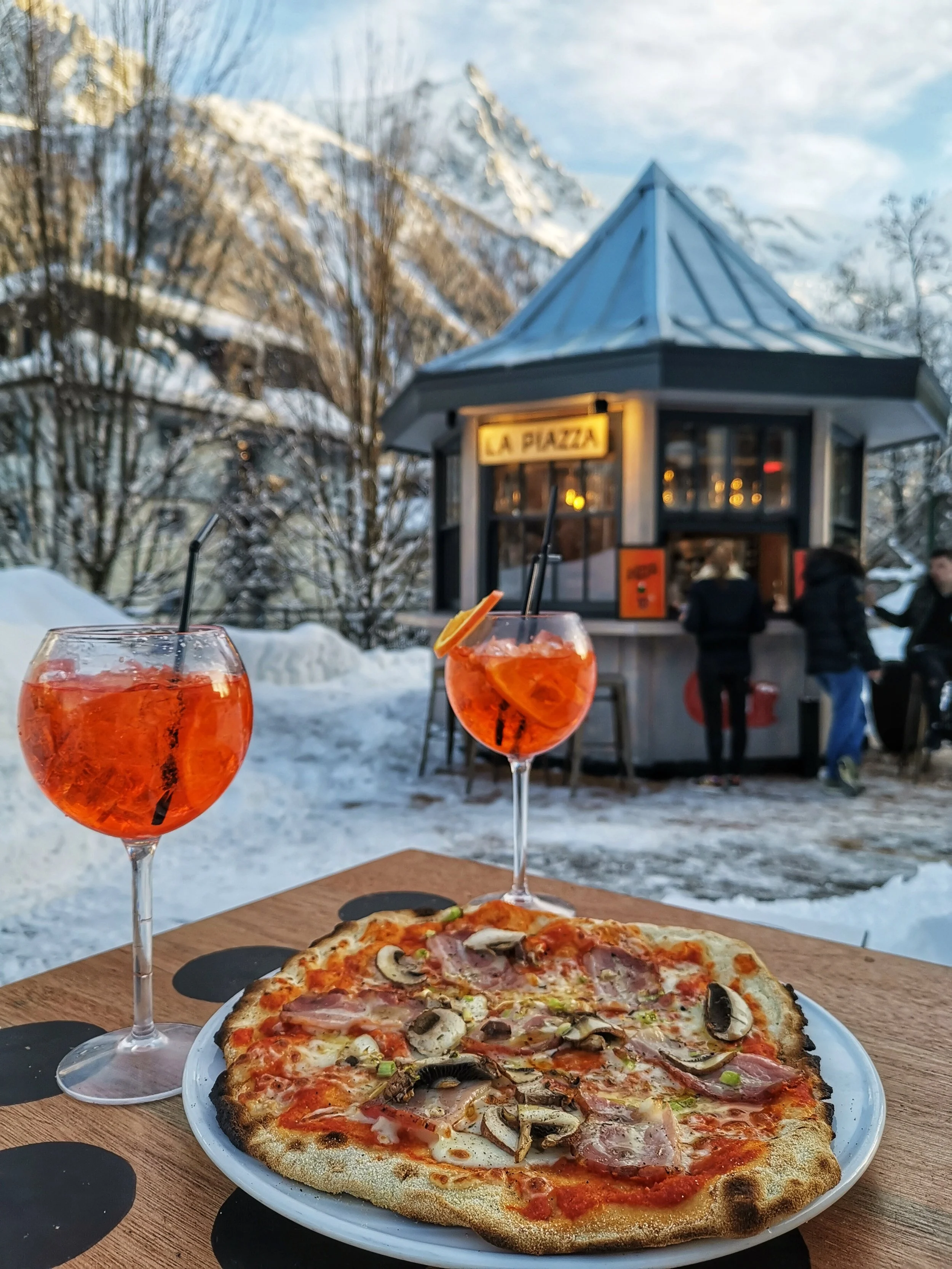 A meal of pizza and two orange cocktails on a wooden table outdoors in a snowy mountain setting, with a small building and people in the background.