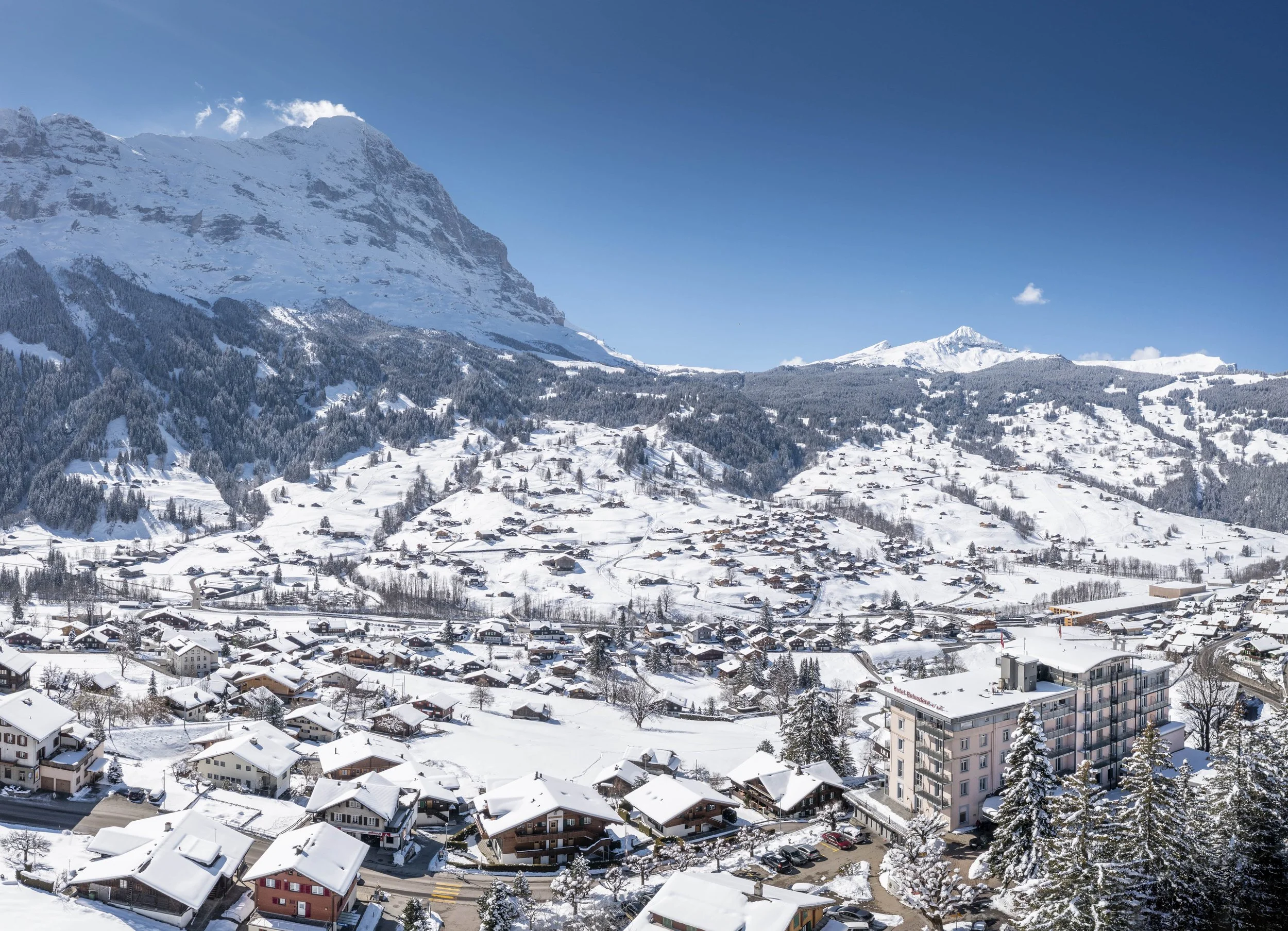 Snow-covered mountain village in a valley with houses and a multi-story building, surrounded by snow-capped mountains and clear blue sky.