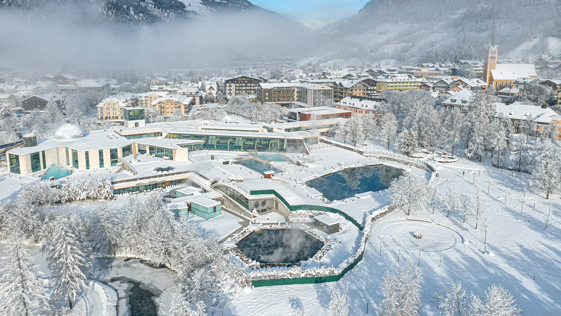 A snowy landscape with a modern spa or resort featuring multiple pools and buildings, surrounded by snow-covered trees and a town with residential buildings and a tall clock tower in the background.