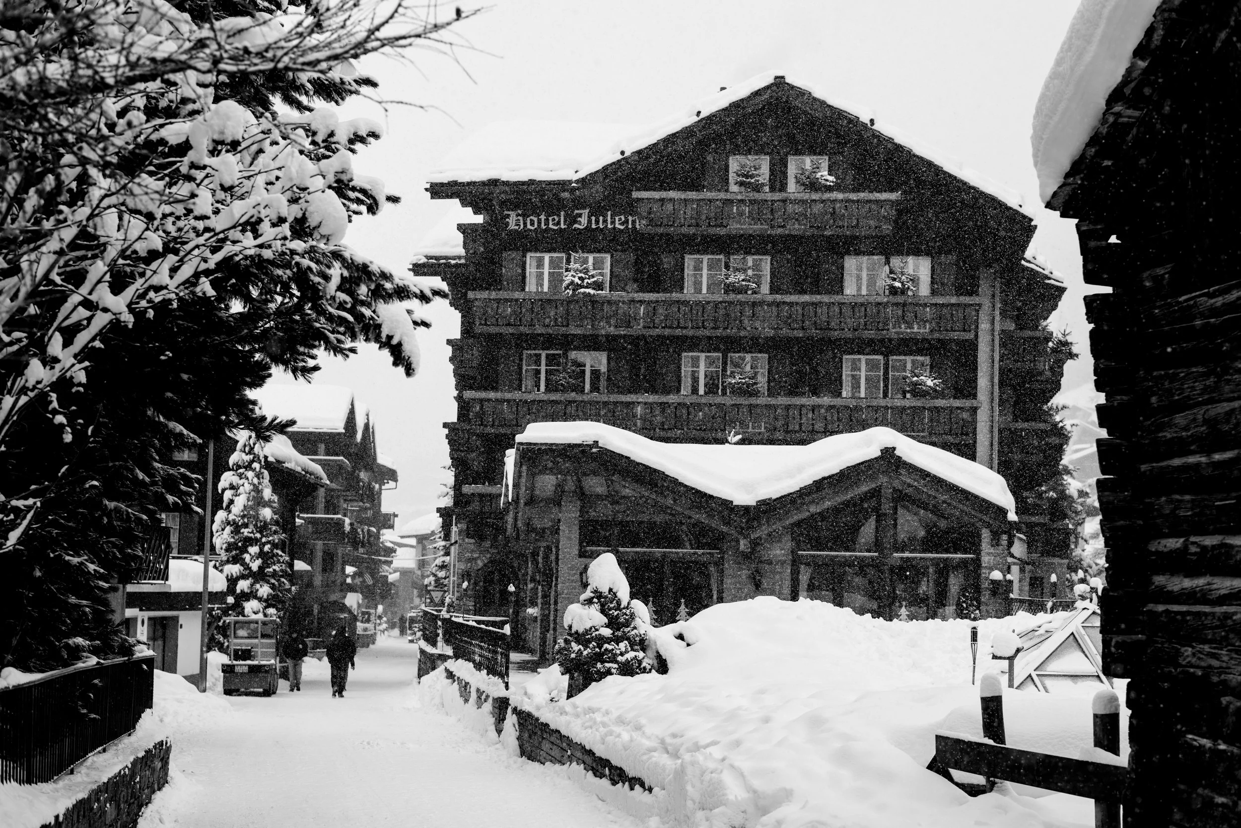 Snow-covered street view with a large wooden hotel named 'Hotel Julen' in the background, surrounded by snow-laden trees and buildings, with people walking along the snow-blanketed sidewalk.