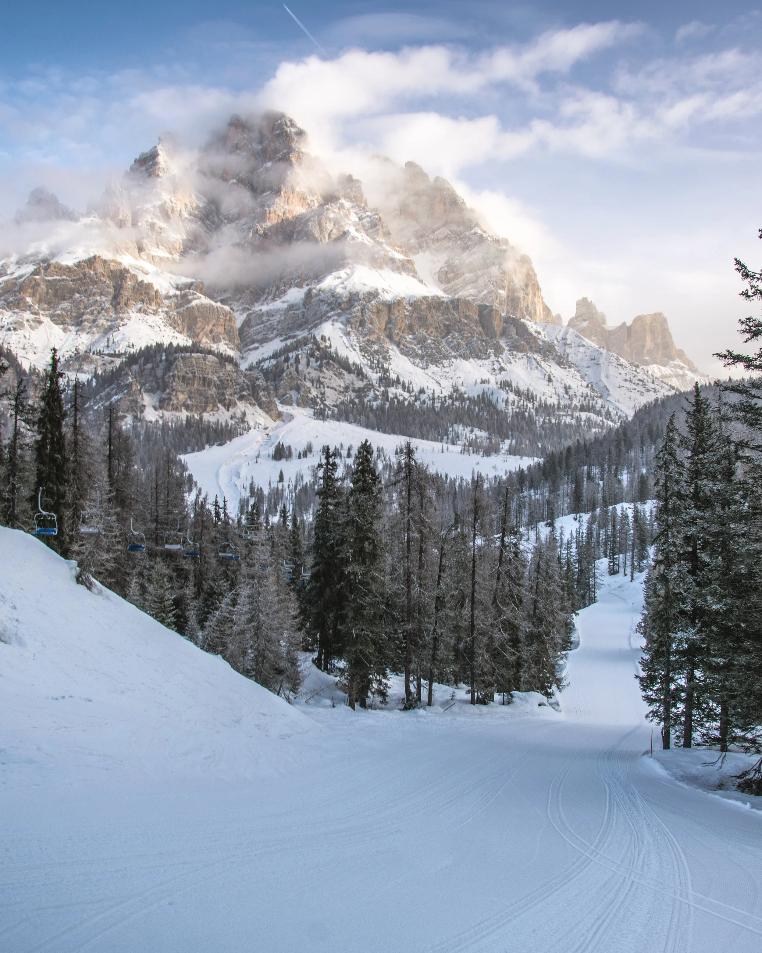 Snow-covered ski slope in a forested mountain landscape with rocky peaks and clouds in the background