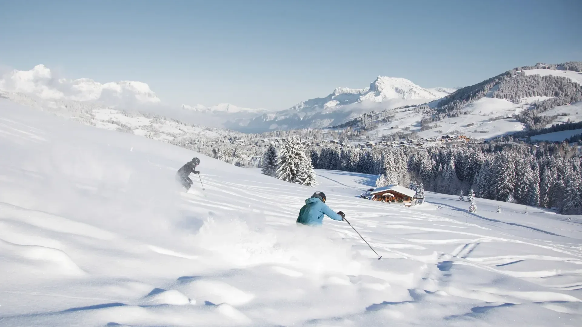 Two skiers in fresh snow cruising down the mountain in blue and black jackets. Sunny weather in Megeve ski resort.