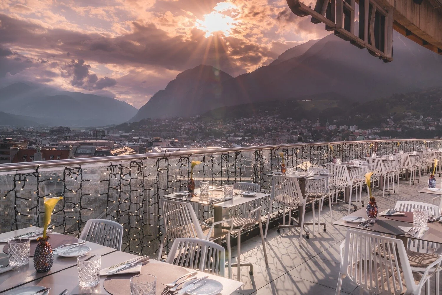 Outdoor restaurant deck with tables set with plates, glasses, and vases with calla lilies, overlooking a city at sunset with mountains in the background.