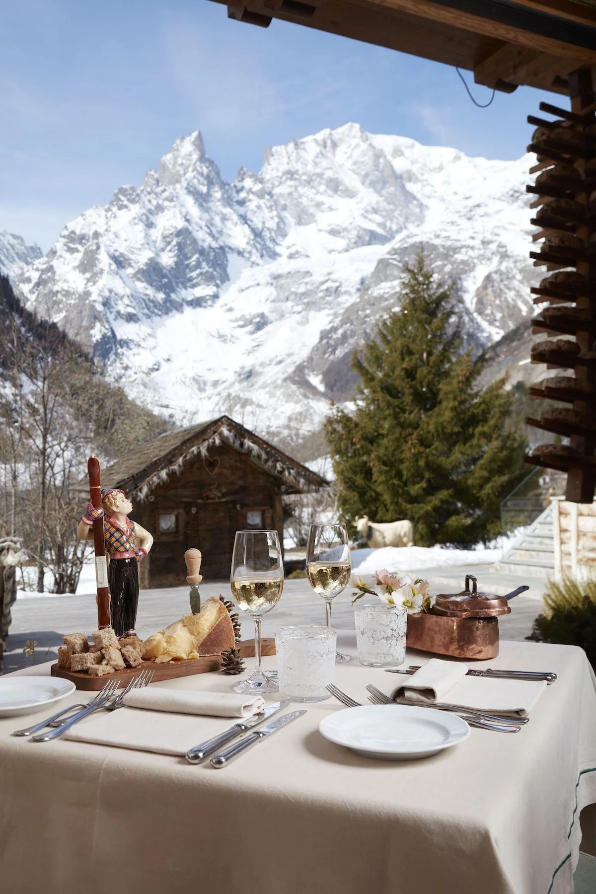 A table set with white plates, silverware, two glasses of white wine, a small flower arrangement, and a copper pan, with a mountain scene of snow-capped peaks, trees, and a wooden cabin in the background.
