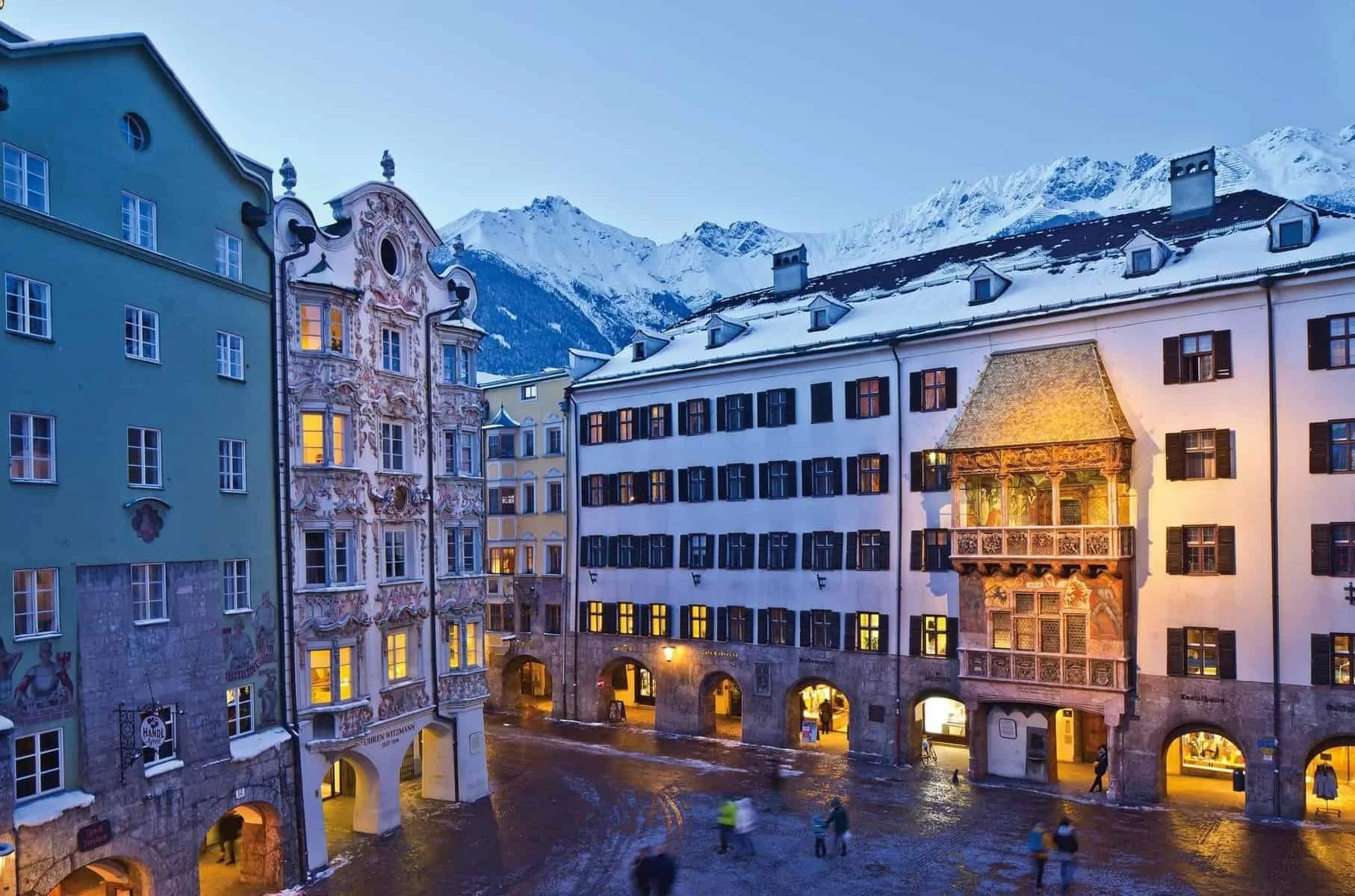 Snow-covered historic buildings on a street in an alpine town with mountains in the background during the evening. Warm lights glow from the windows.