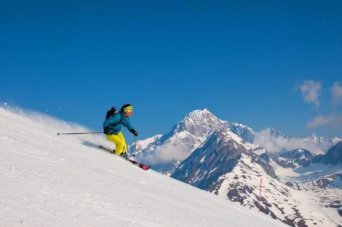 Skier with yellow trousers and blue jacket skiing down the mountain. Fresh snow and blue skies with a peak in the background.