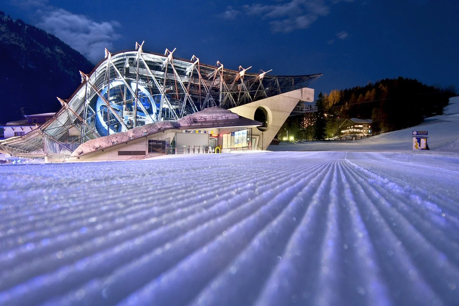 Night view of a modern ski jump facility with snow groomed surface, illuminated by lights, mountains in the background, and trees on the side.
