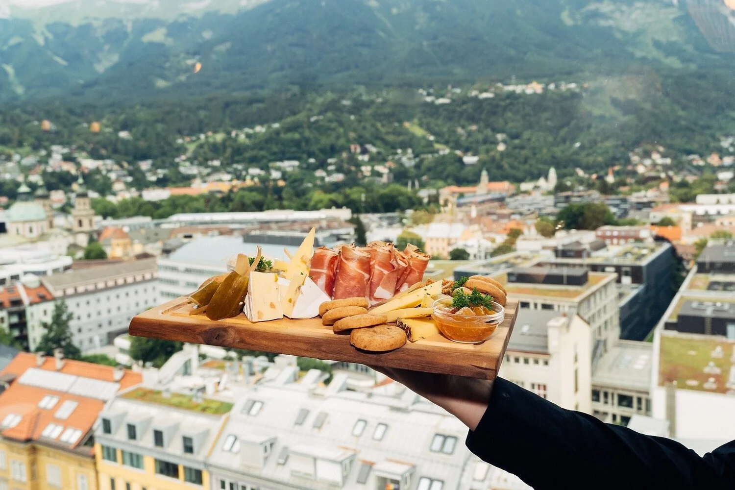 Person holding a wooden cheese and charcuterie board with assorted cheeses, cured meats, crackers, pickles, and honey, with a cityscape and mountains in the background.