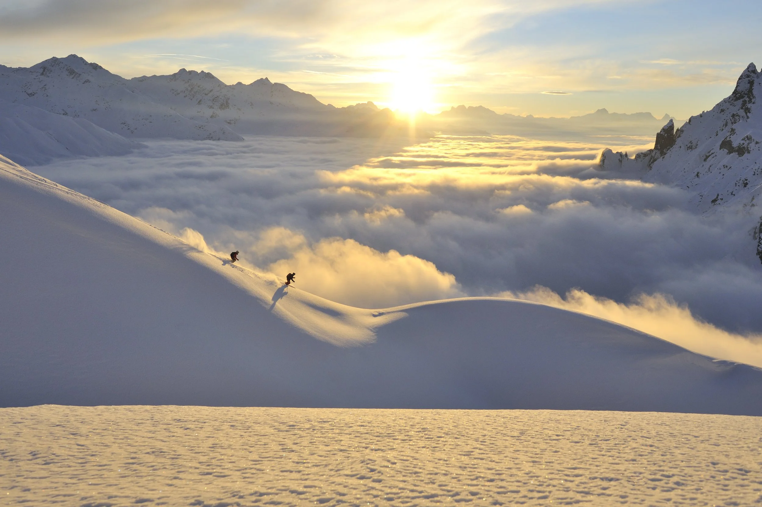 Two skiers descending a snow-covered mountain slope at sunset with mountains and clouds in the background.