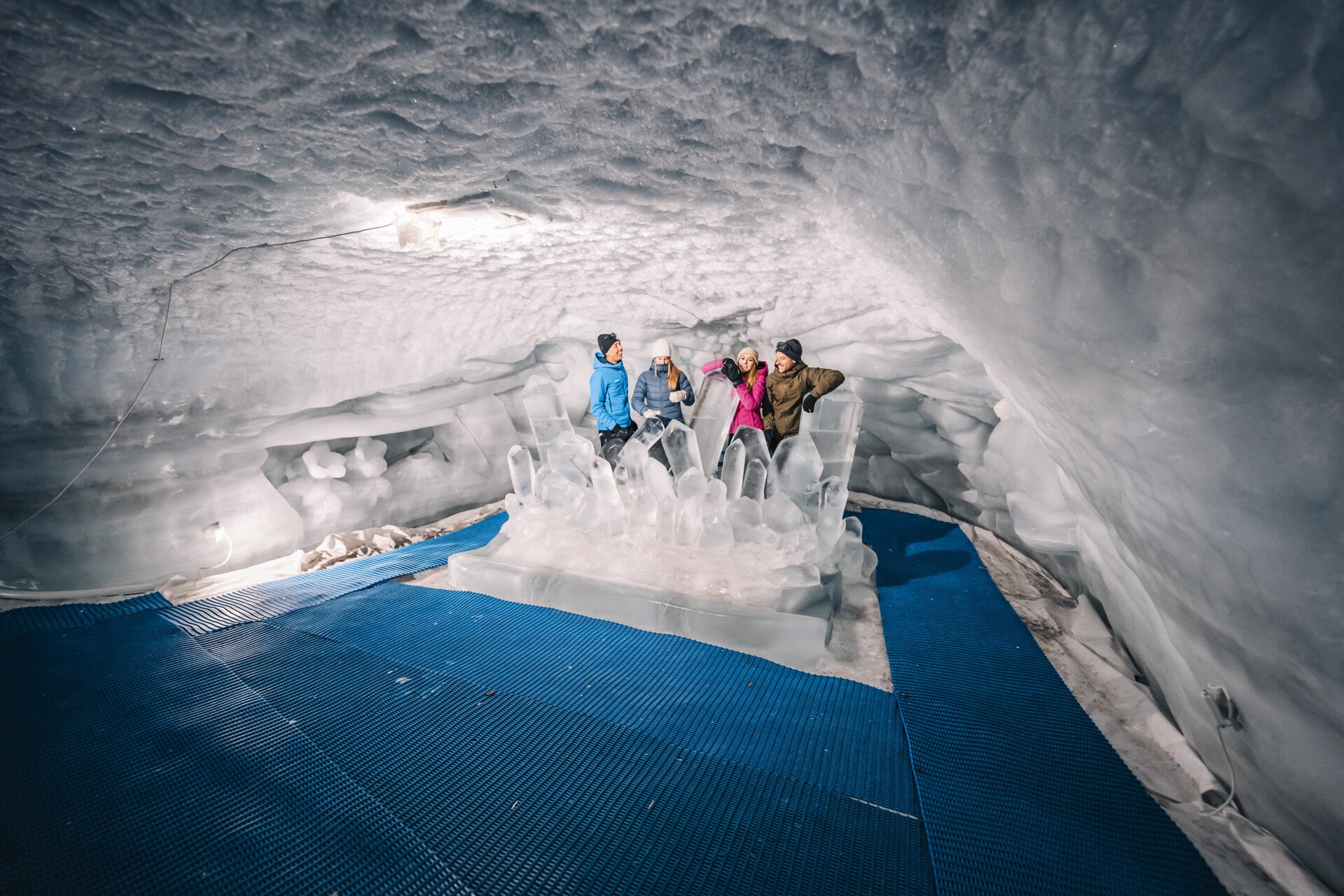Four people stand inside an ice cave, overlooking a large ice sculpture with tall, pointed ice formations. The cave walls are made of ice and snow, and the floor has blue matting.