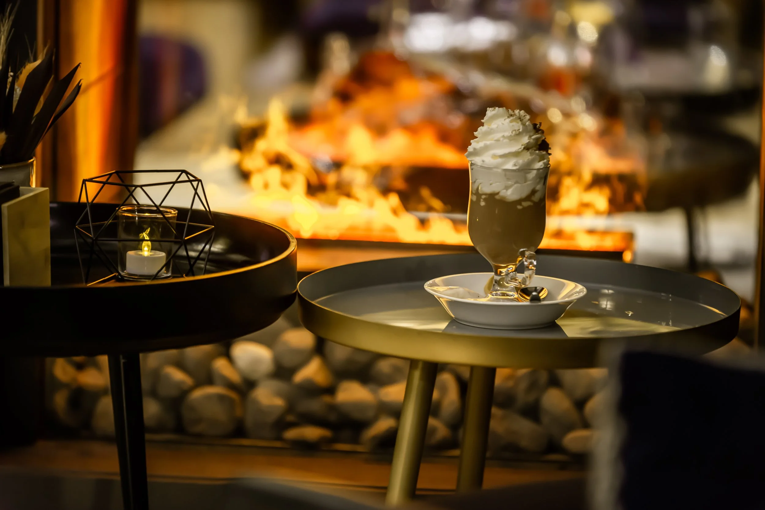 A tall glass of hot chocolate, whipped cream-topped beverage on a white saucer with spoon, on a small round black table near a fireplace.