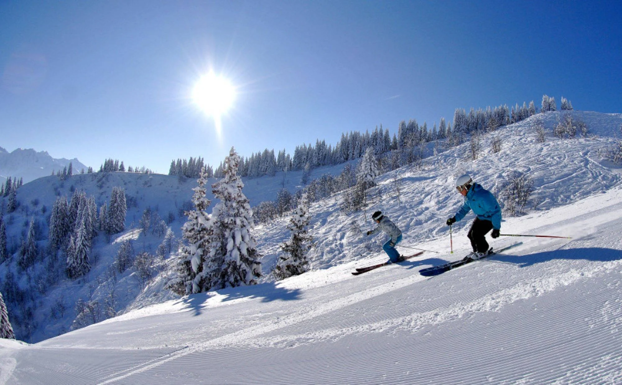 Two individuals skiing down a sunny piste with fresh snowfall and perfect groomed runs.