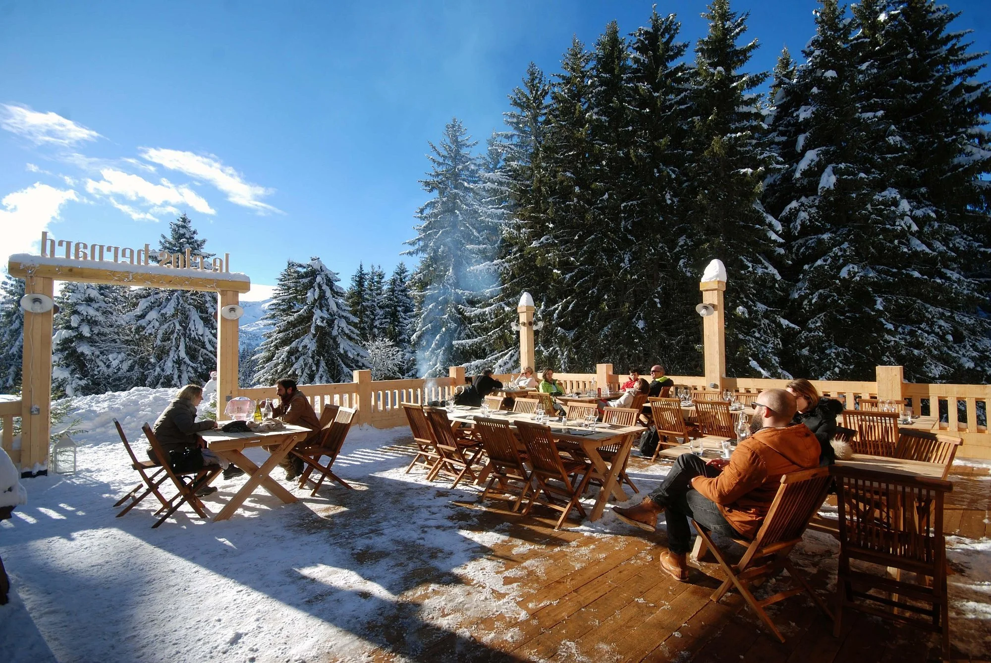 Clos Bernard, Meribel terrace. People enjoying drinks in the sun amidst the snowy pine trees.