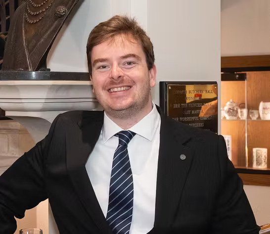 A young man in a black suit and tie smiling at a social gathering.
