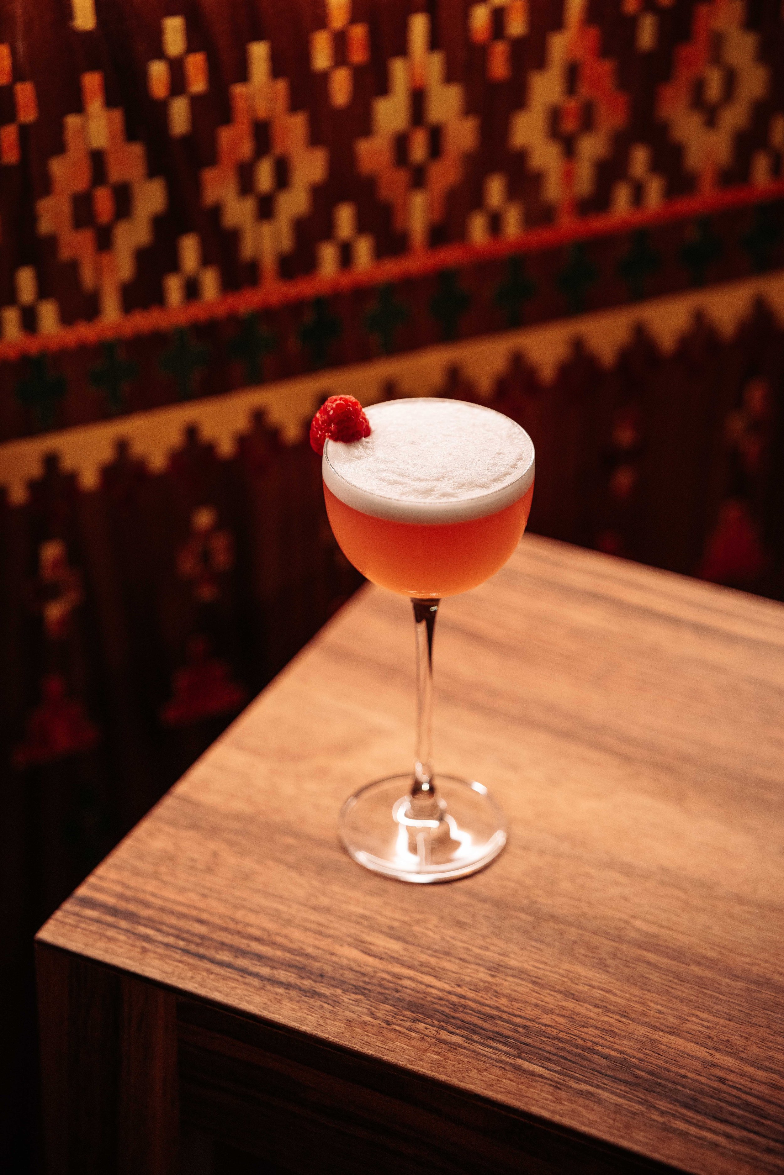 A cocktail in a coupe glass with a frothy top and a red berry garnish, placed on a wooden table.