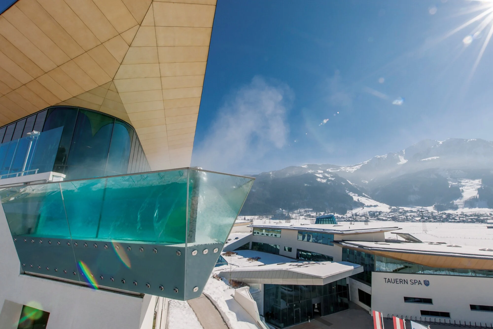 Modern spa building with large glass windows and balconies, snow-covered landscape in the background, mountains, clear blue sky, and bright sunlight.