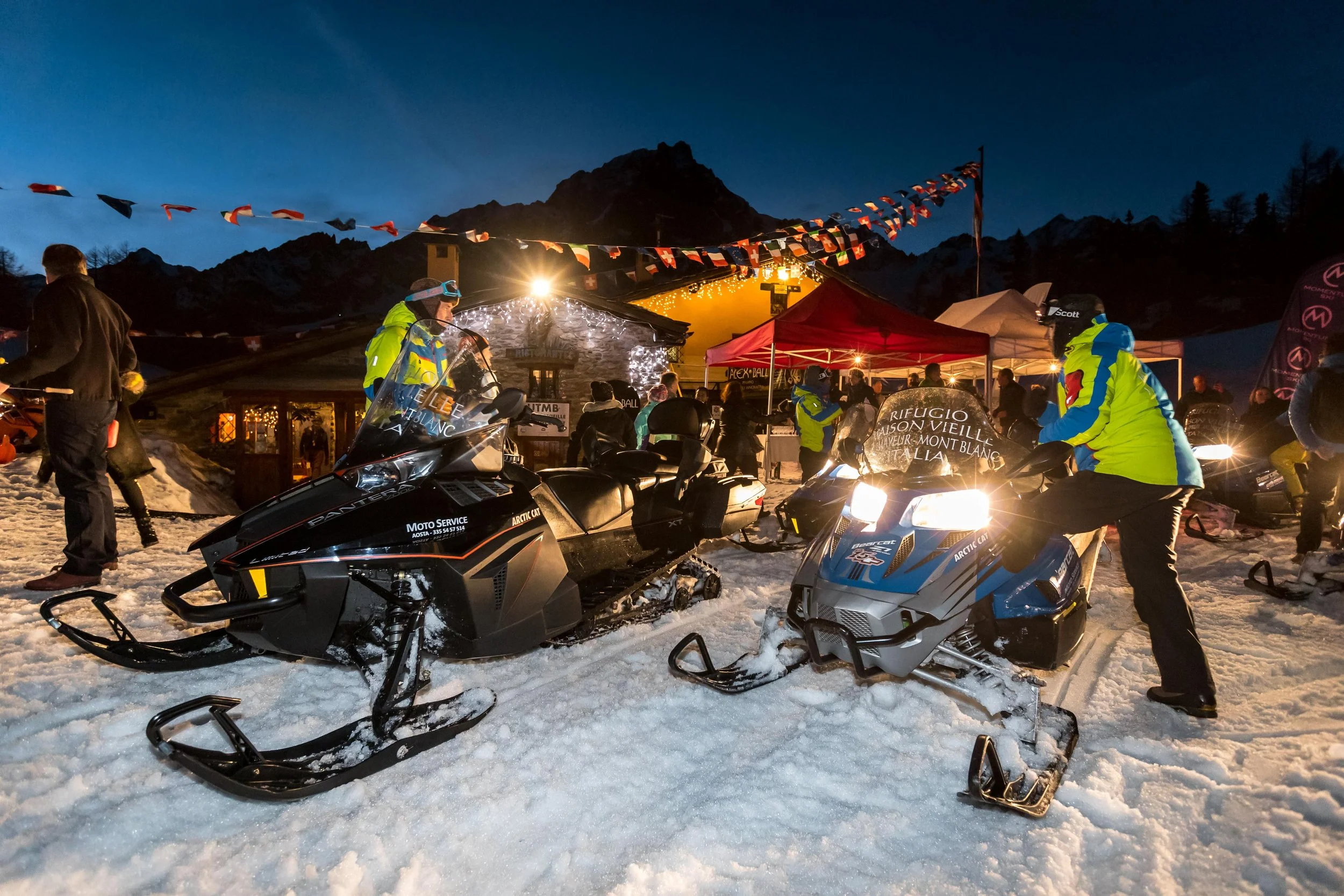 People in winter clothing and helmets at night in snow-covered mountain area, with snowmobiles, tents, and festive lights.