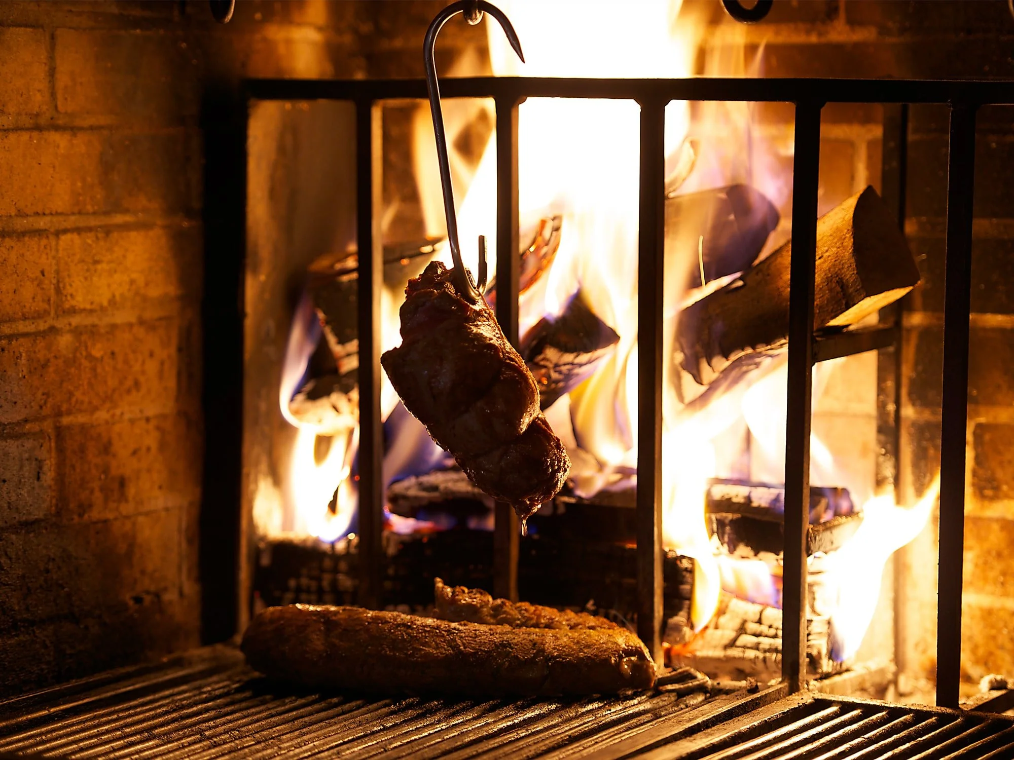 A skewer with meat cooking in a fireplace with a metal grate and flames in the background.