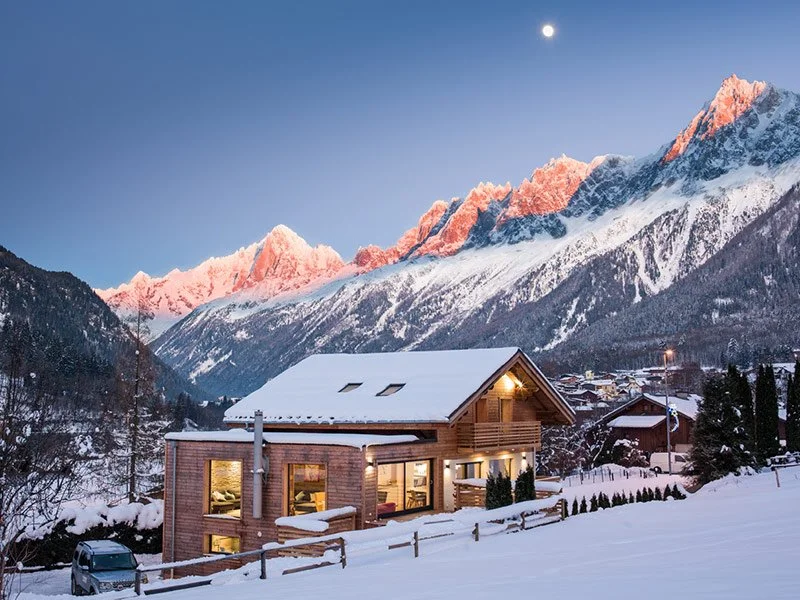 Snow-covered mountain landscape at sunset with a wooden house in the foreground.