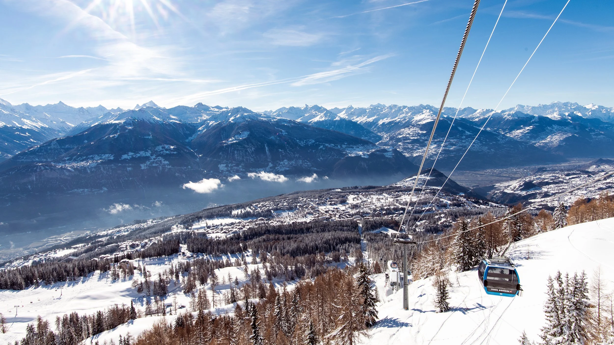 Snow-covered mountains and trees with a ski lift in the foreground under a clear blue sky.