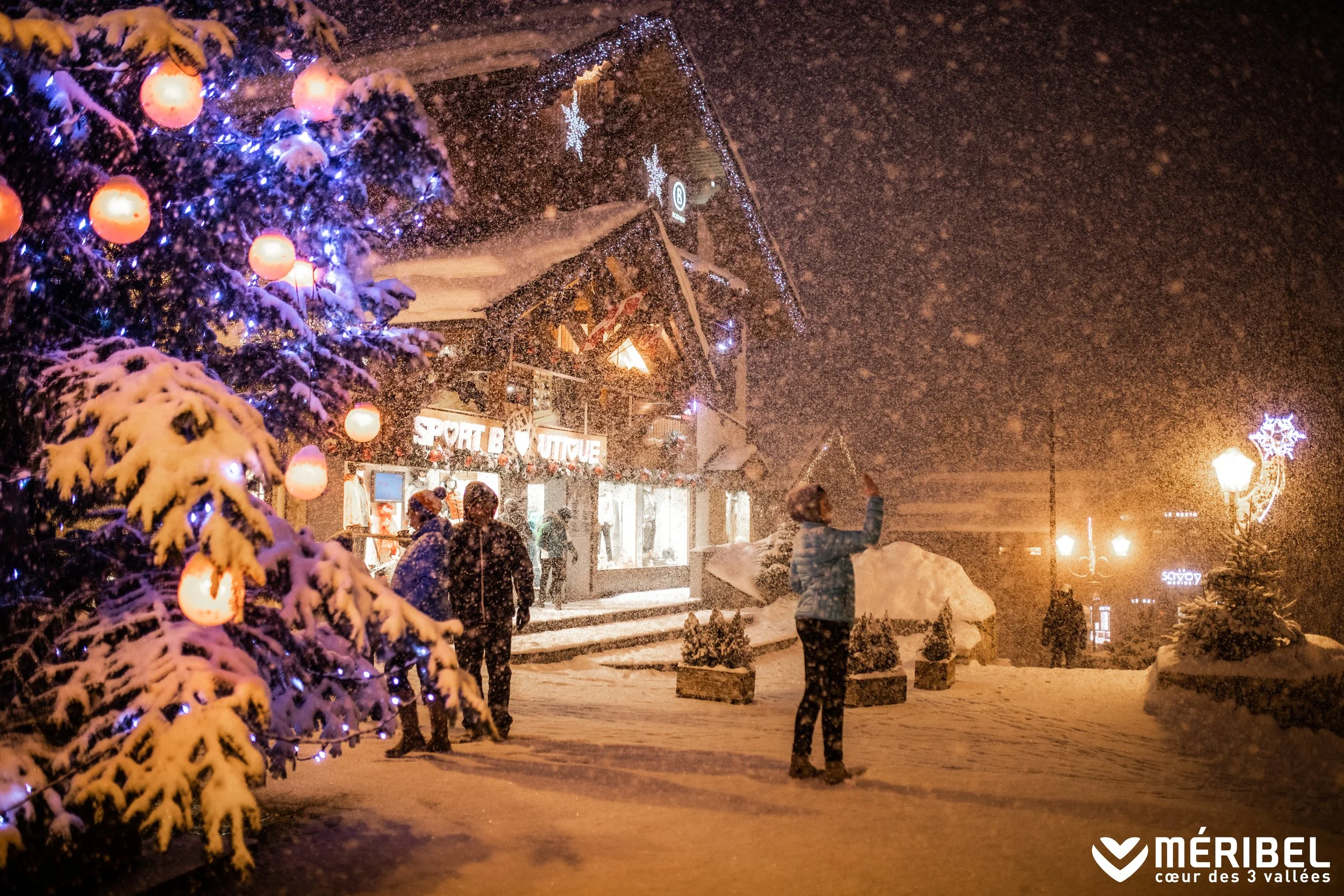 People walking in a snow-covered outdoor shopping area decorated with Christmas lights and ornaments at night, with a snow-covered tree in the foreground and a shop sign that reads 'SPORT B' in the background.
