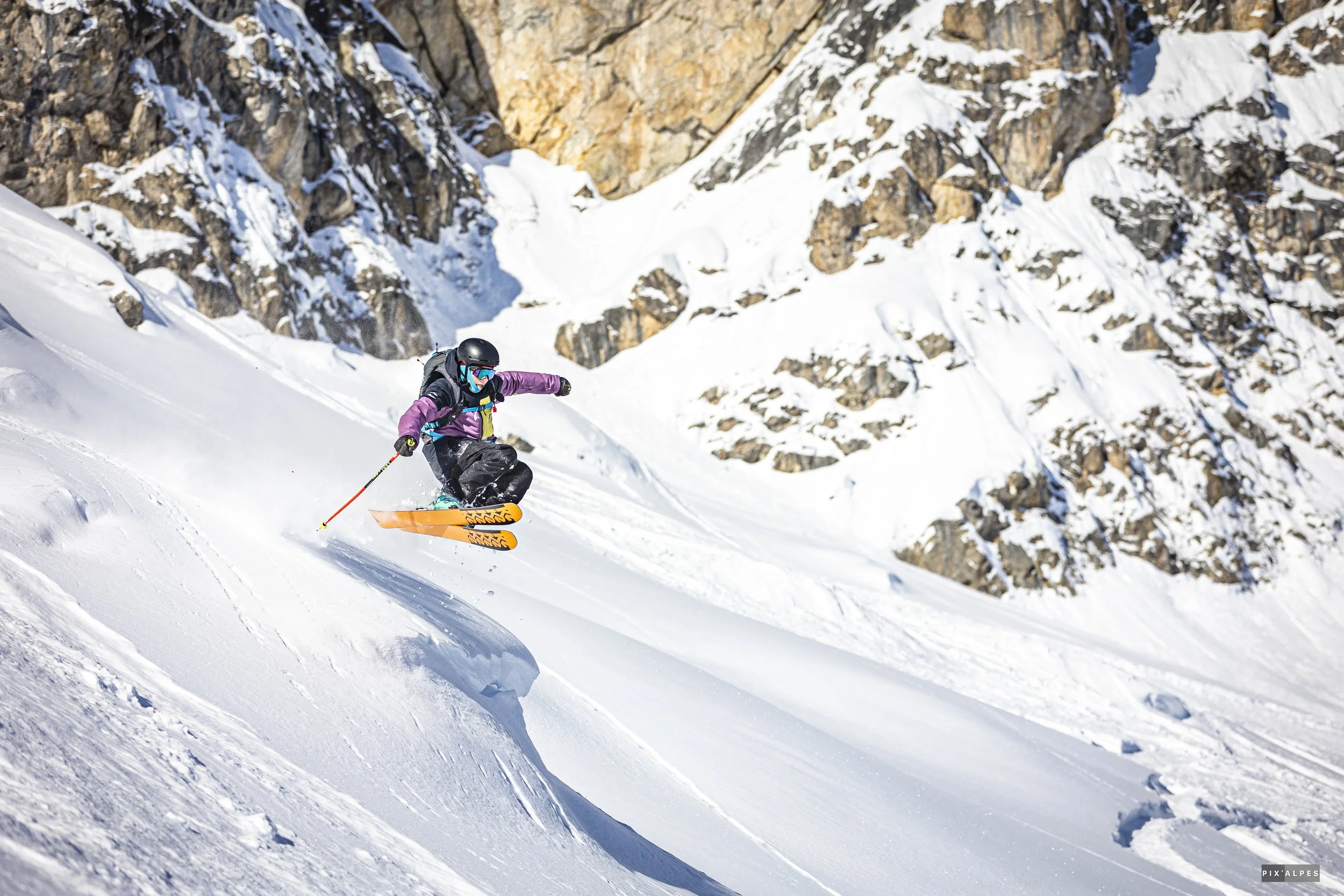 A skier in purple and black gear navigating down a snow-covered mountain slope with rocky cliffs in the background.