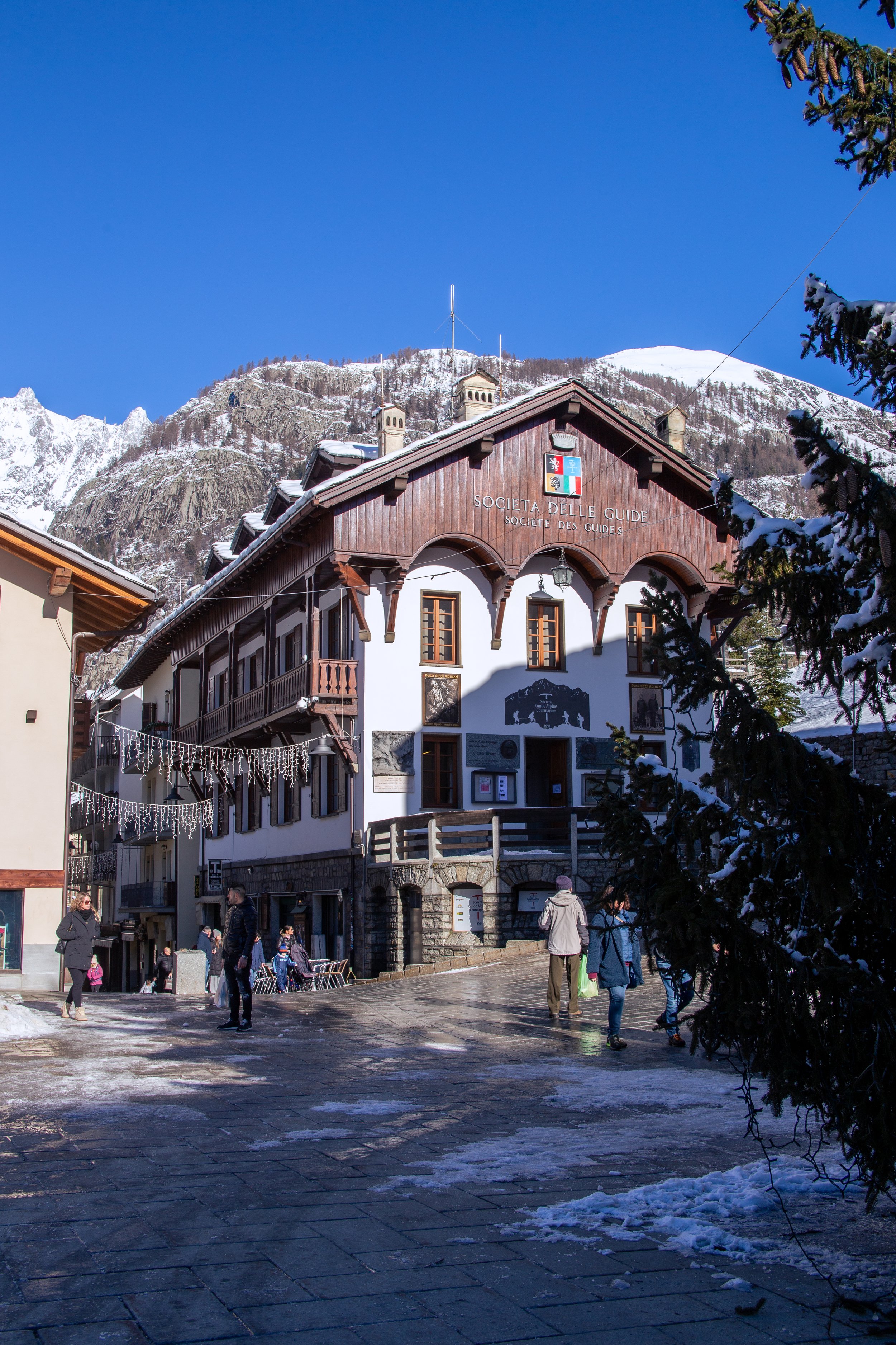 A snowy street scene in a mountain village with a prominent wooden building labeled "Societa Delle Guide" and "Société des Guides" on the front, surrounded by snow-covered mountains and a clear blue sky.