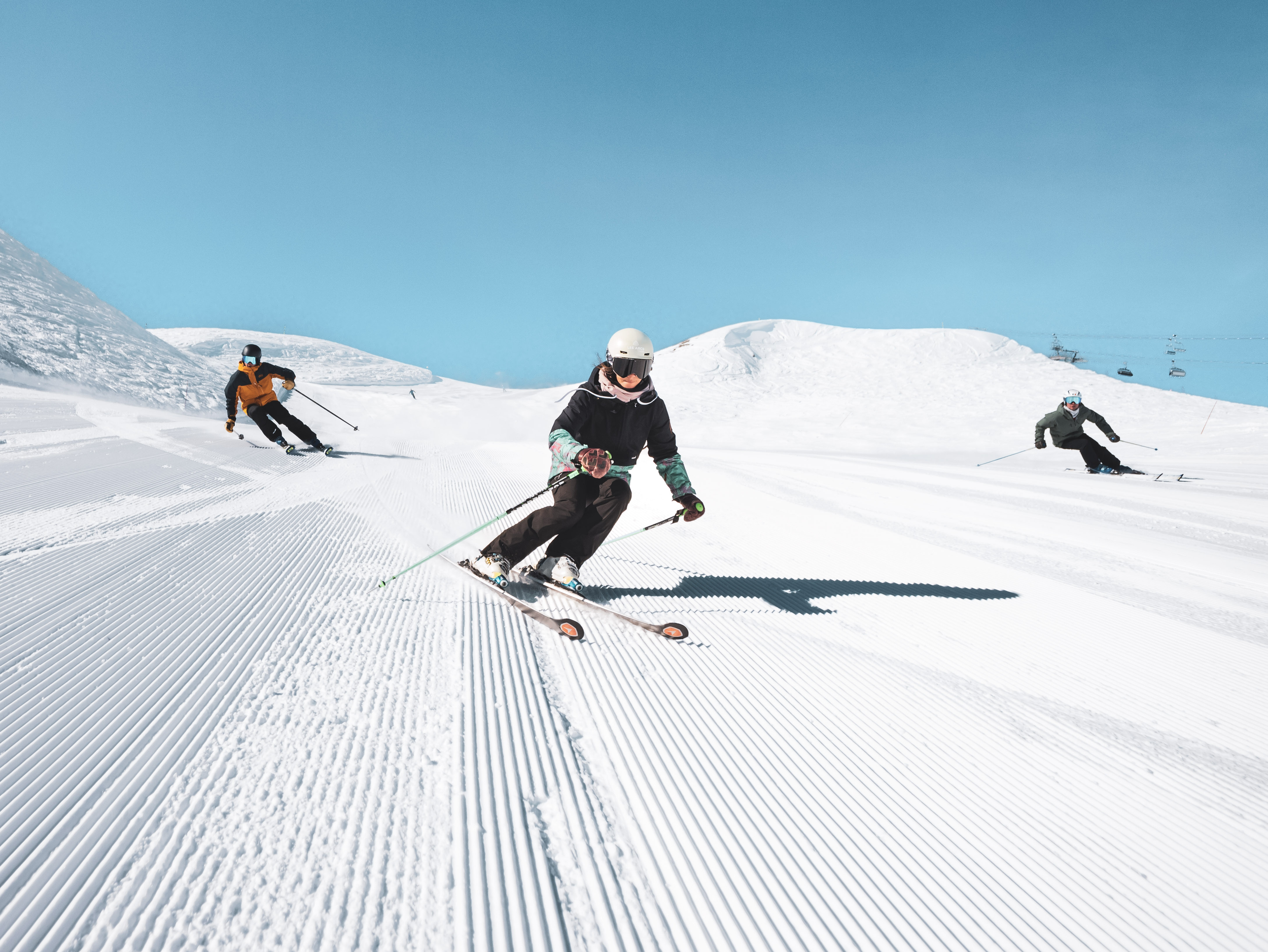 Three skiers skiing down a groomed snow slope on a clear day, with ski lifts visible in the background.