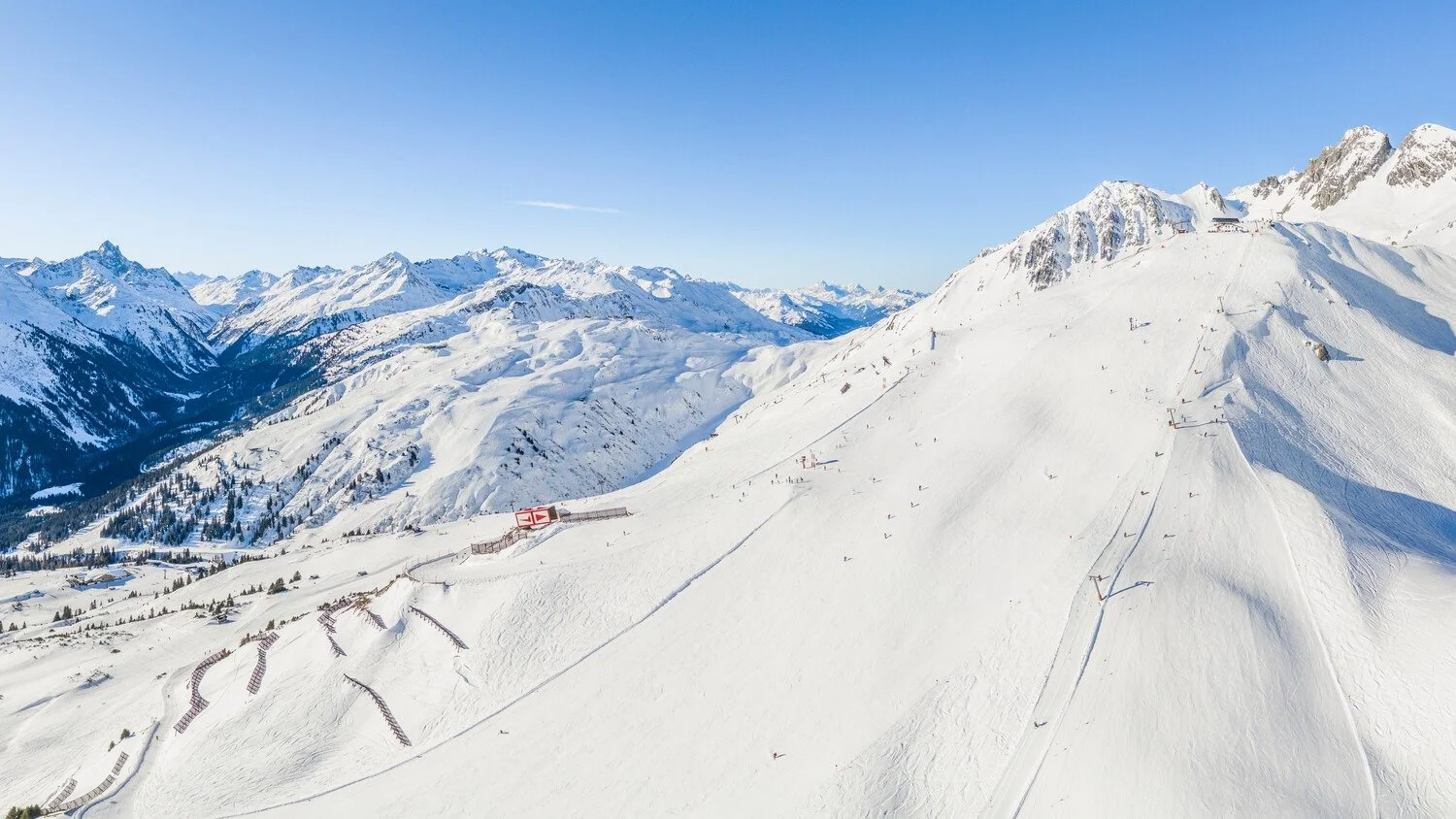 Snow-covered mountain landscape with ski slopes and chairlifts, and distant mountain peaks under a clear blue sky.