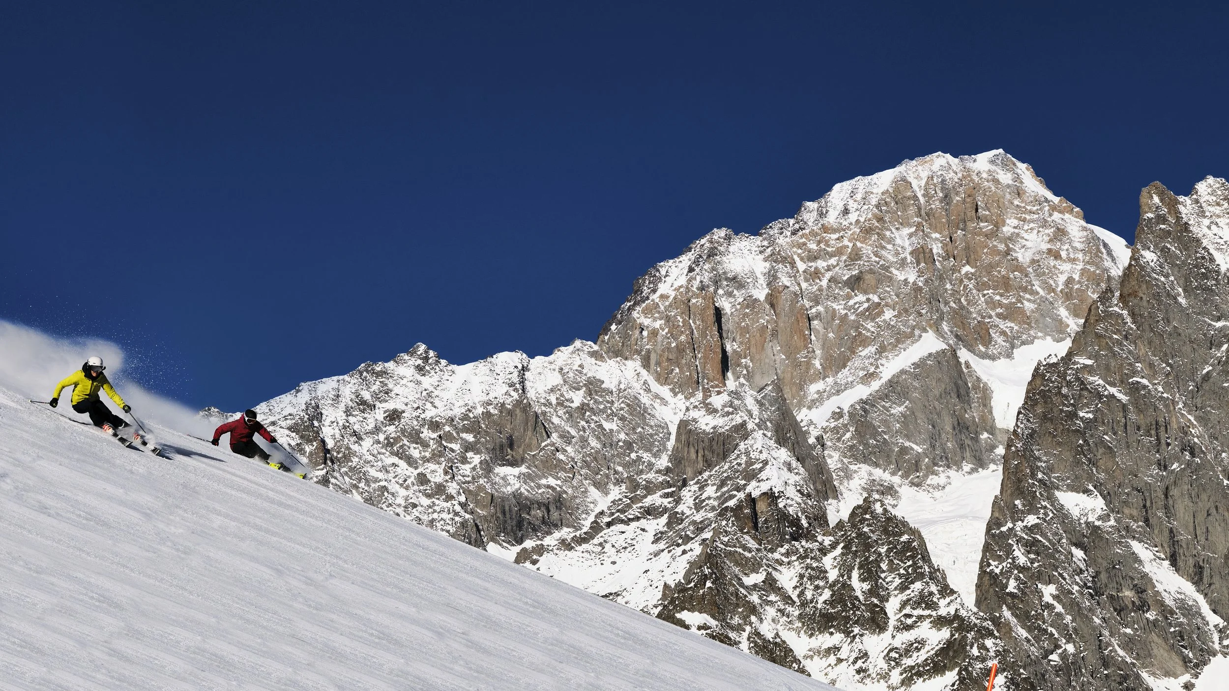 Two skiers carving down the mountain on a fresh blue sky day and fresh snowfall.
