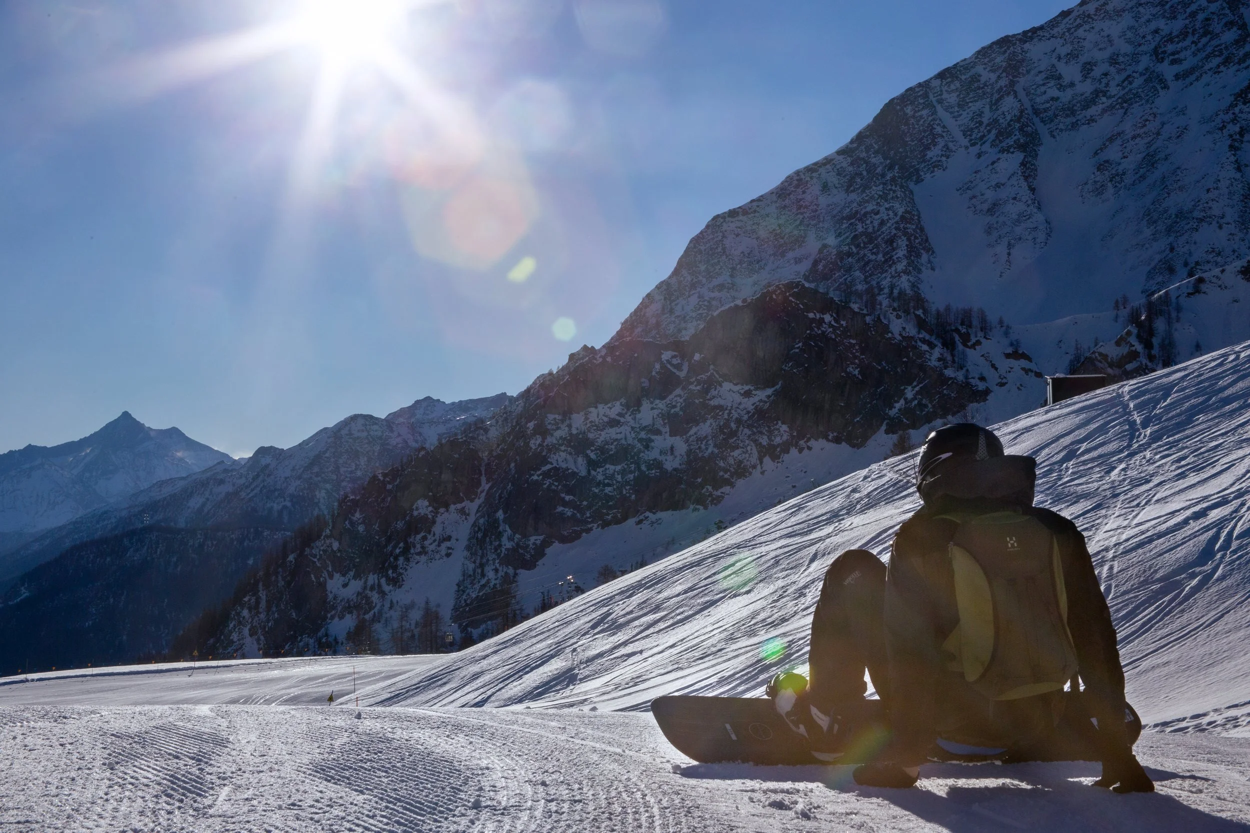 A person in winter gear and a helmet sits on snow with a snowboard, looking towards snow-covered mountains under a bright sun in a clear blue sky.